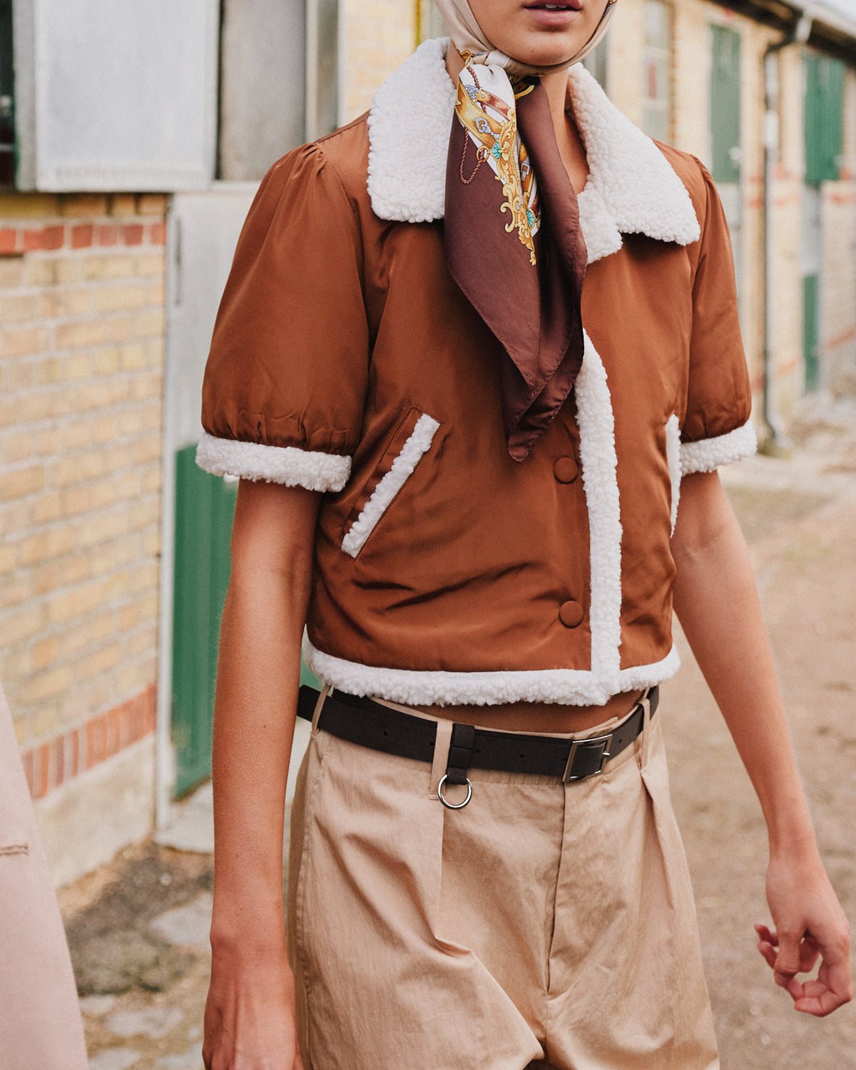 Campagne-close-up van een model met een roestbruine jas met witte shearling-afwerking en een zijden sjaal met dessin.