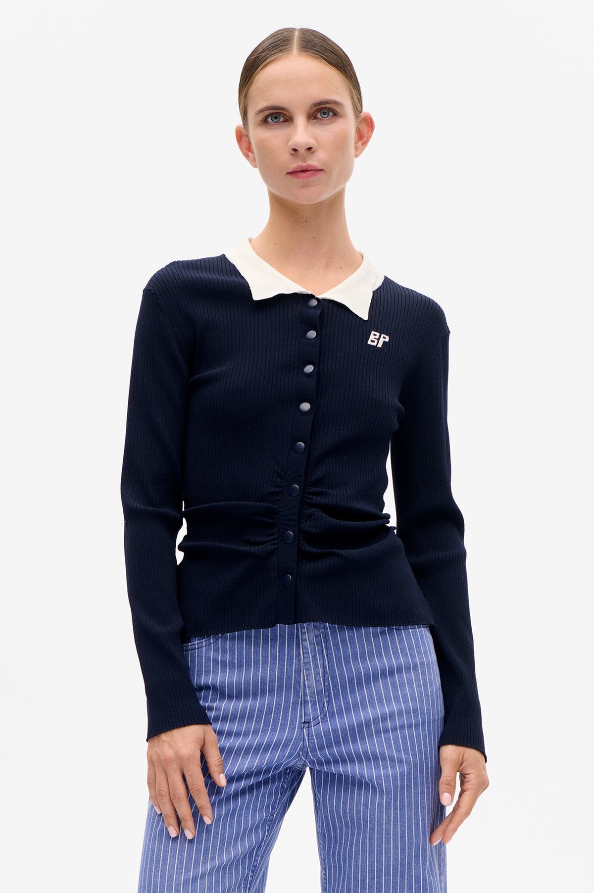 Front view of a navy ribbed button-front cardigan with a white pointed collar and small chest logo, paired with blue pinstripe trousers.