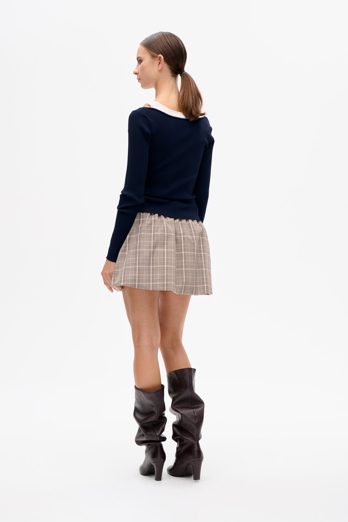 Model shown from the side and back in a navy ribbed long-sleeve top with a white collar and beige plaid mini skirt.
