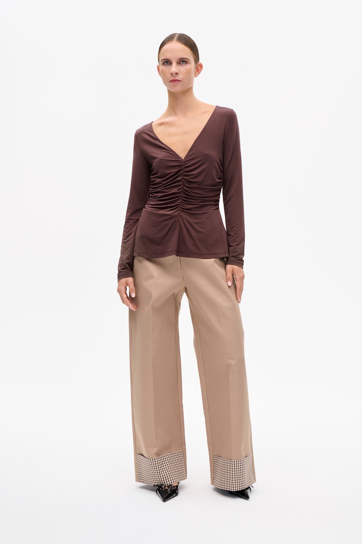 Model trägt ein pflaumenfarbenes, langärmliges Top mit V‑Ausschnitt und geraffter Vorderseite sowie eine beige, weitgeschnittene Hose mit karierten Bündchen.