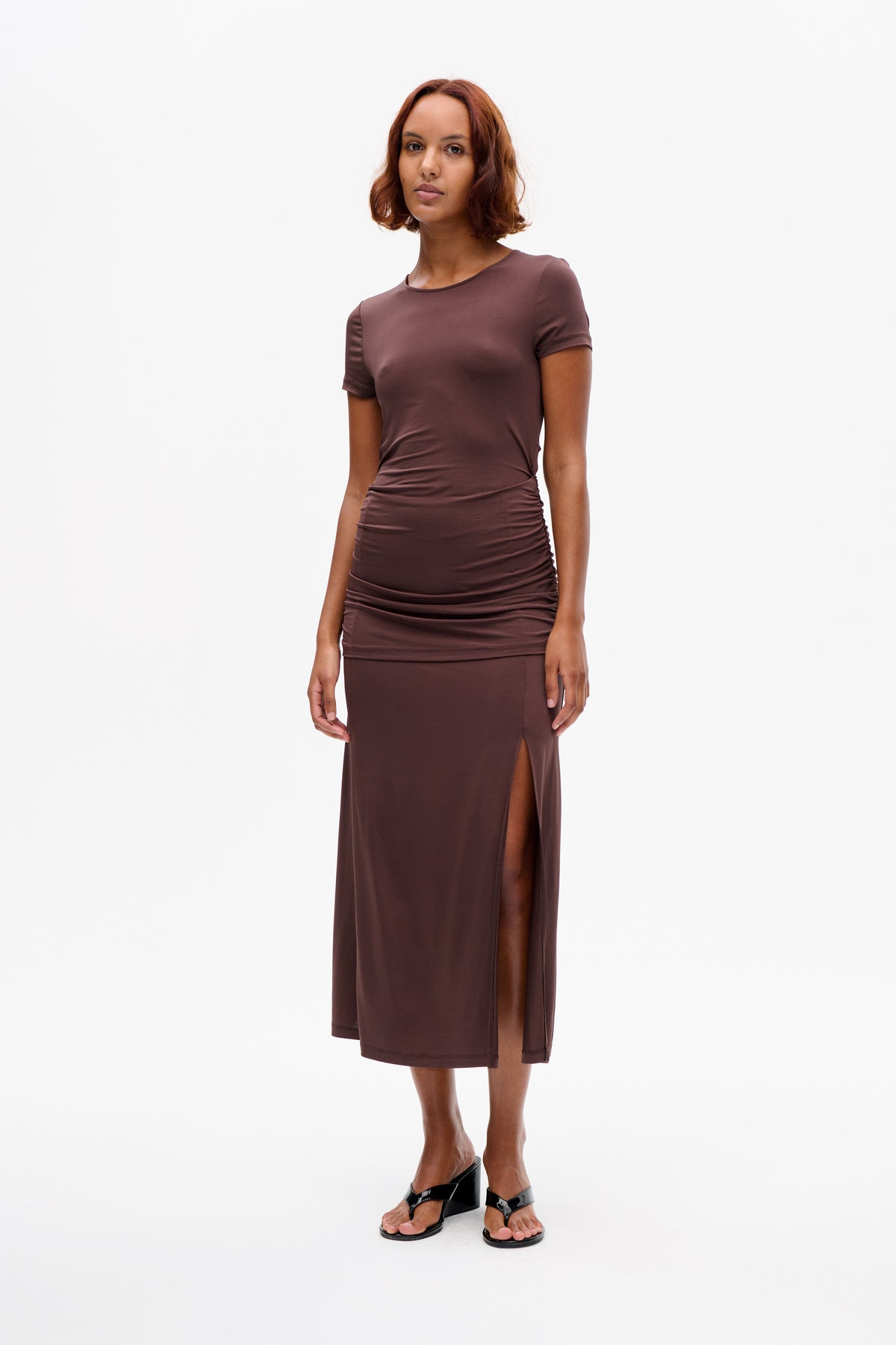 Front view of a model wearing a dark mauve short-sleeved midi dress with a round neckline, ruched waist and side slit.