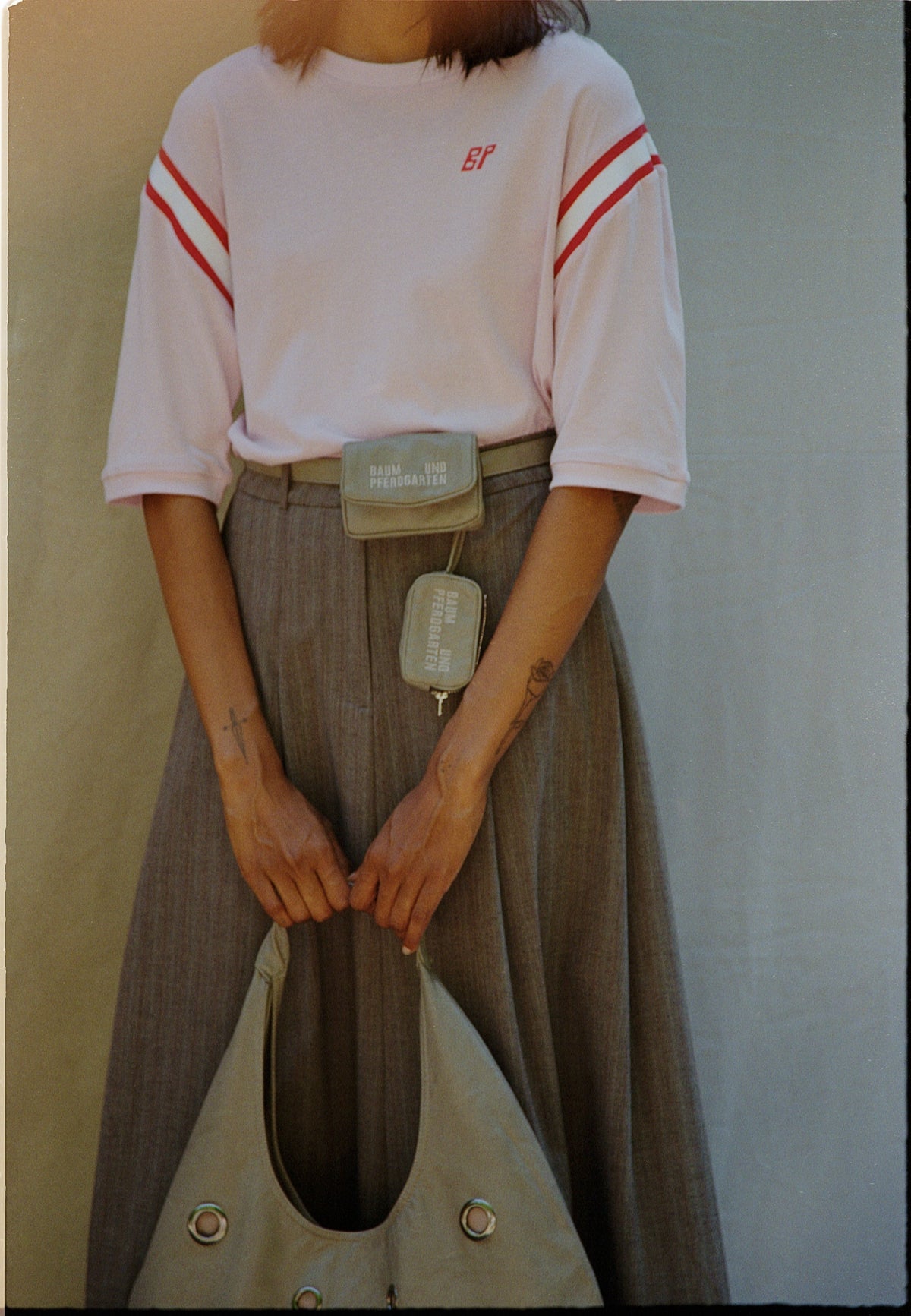 Beschnittenes Kampagnenporträt eines Models in weißem Sweatshirt mit roten Ärmelstreifen und grauen Hosen, das eine kleine Crossbody‑Tasche und eine große salbeigrüne Tote trägt.
