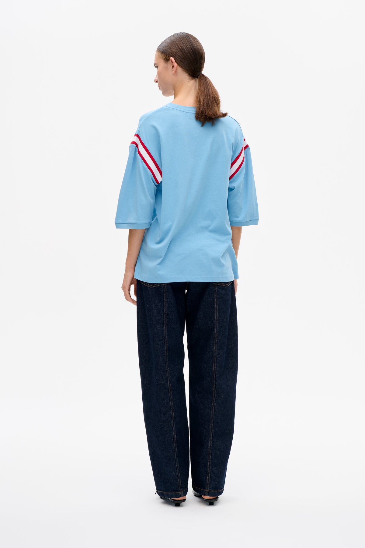 Rückenansicht eines Models, das ein hellblaues oversized T-Shirt mit rot-weißen diagonalen Streifen an den Ärmeln und dunkle Wide-Leg-Jeans trägt.