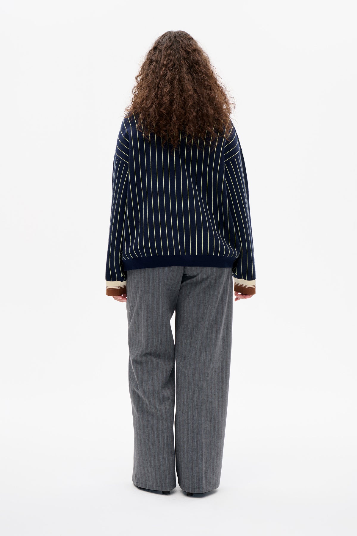 Back view of a model wearing a navy long-sleeved sweater with thin yellow vertical stripes and cream-and-brown striped cuffs.