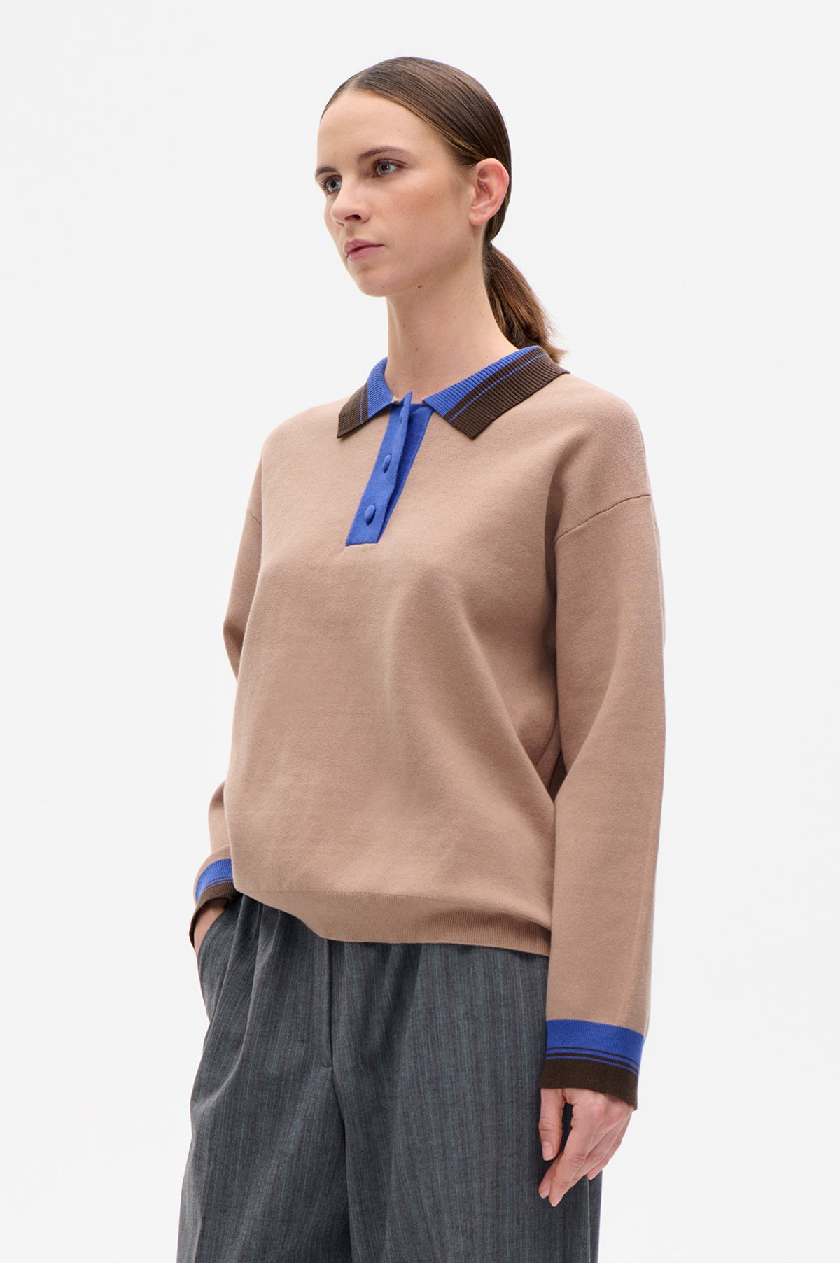 Model wearing a beige long-sleeve knit polo with a bright blue button placket and blue-and-brown striped collar and cuffs