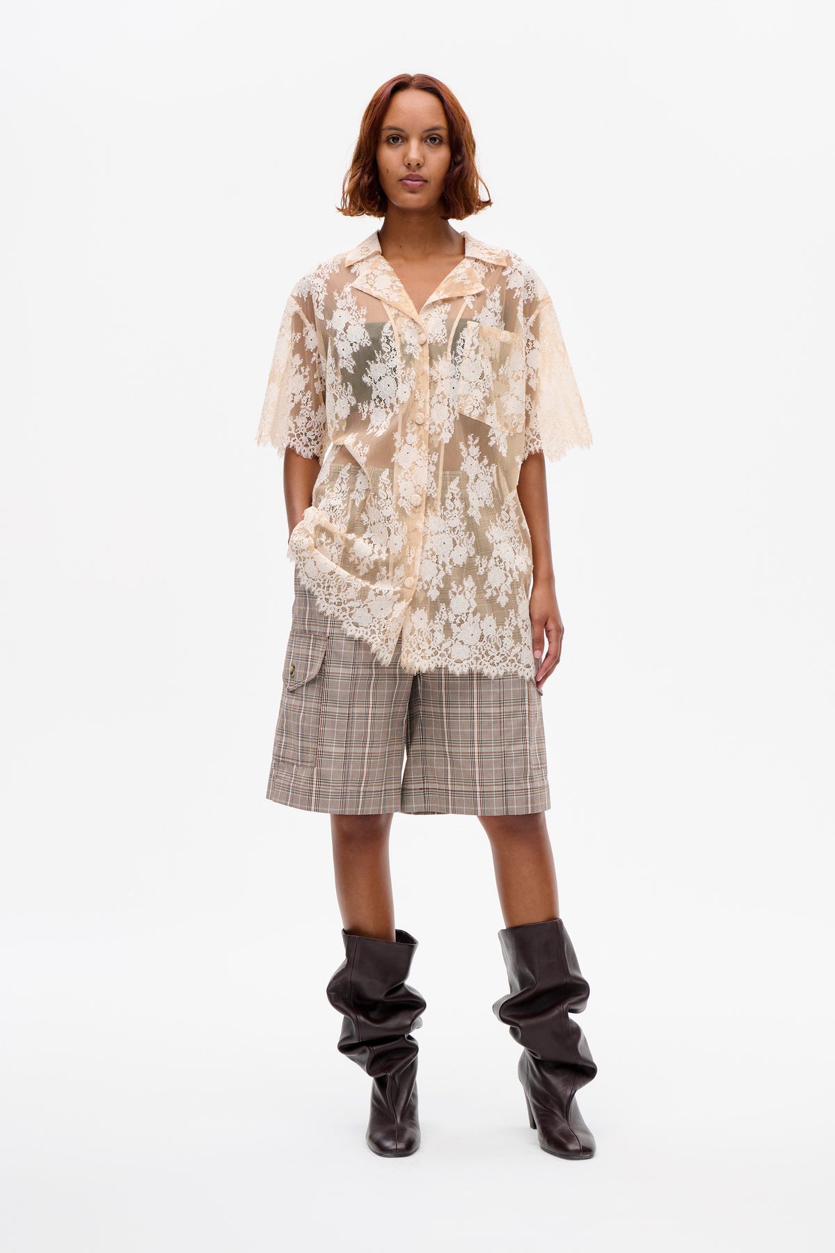 Vorderansicht eines Models, das eine transparente cremefarbene Bluse aus floraler Spitze mit Knopfleiste, beige karierte Shorts und dunkelbraune Slouch-Boots trägt.
