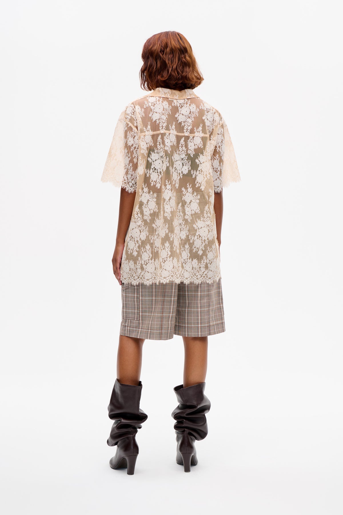Rückenansicht eines Models in einer transparenten, cremefarbenen Tunika aus floraler Spitze mit Kragen, braunen karierten Shorts und dunklen, lässig sitzenden Stiefeln mit Absatz.