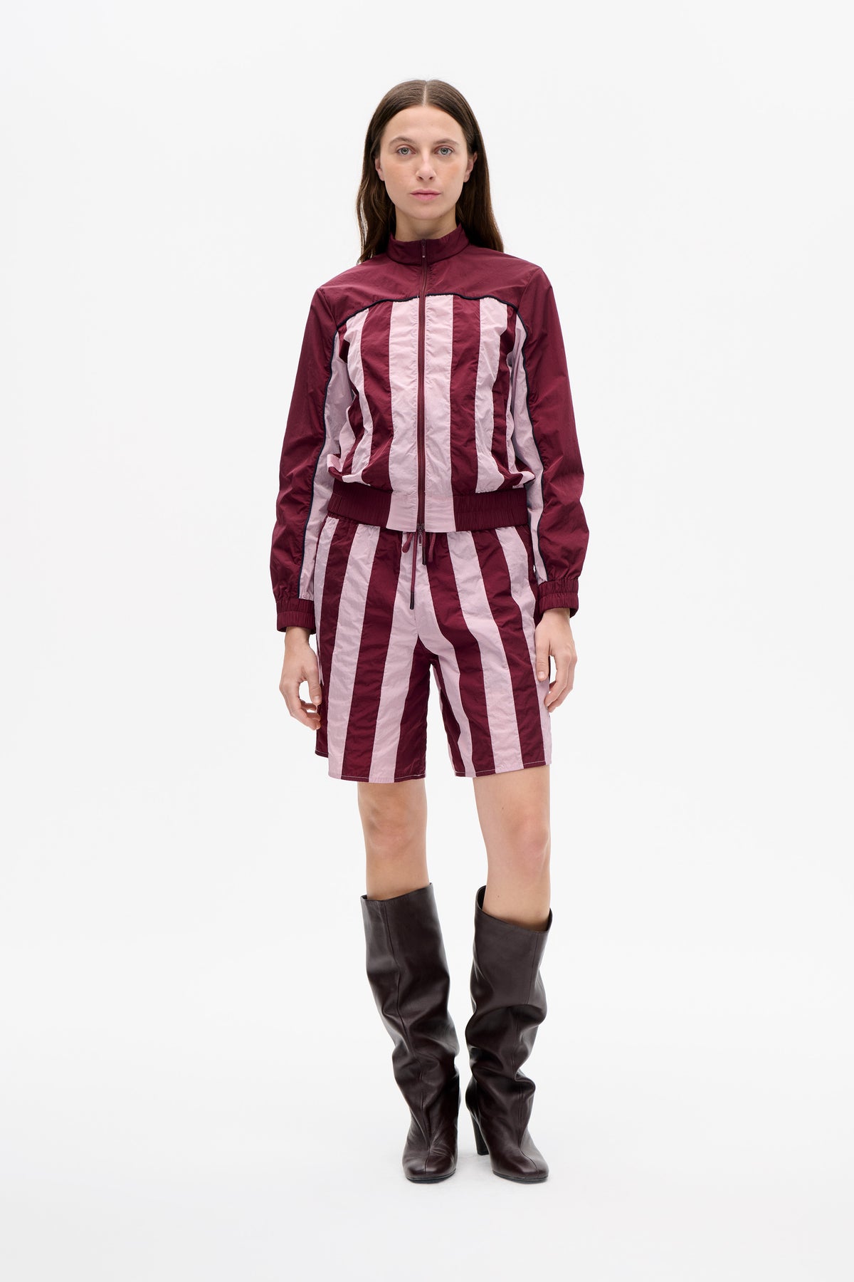 Front-facing model wearing a burgundy and pale pink striped zip-up jacket with matching shorts and dark knee-high boots.