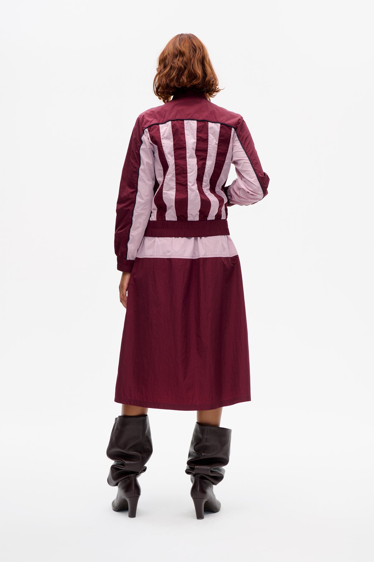 Back view of a model wearing a burgundy jacket with pale pink vertical panels and dark piping, over a matching midi skirt.
