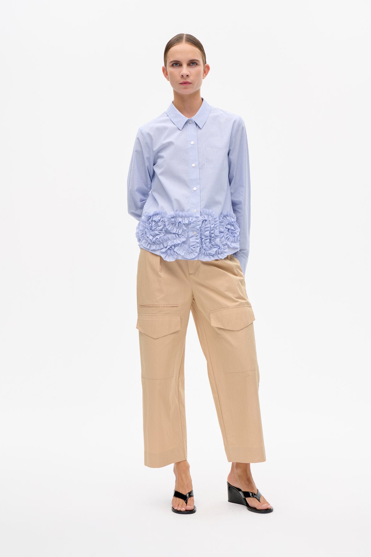 Modell trägt eine hellblau-gestreifte Hemdbluse mit Rüschensaum, sandfarbene, gekürzte, weitgeschnittene Cargo-Hose und schwarze Keilsandalen.