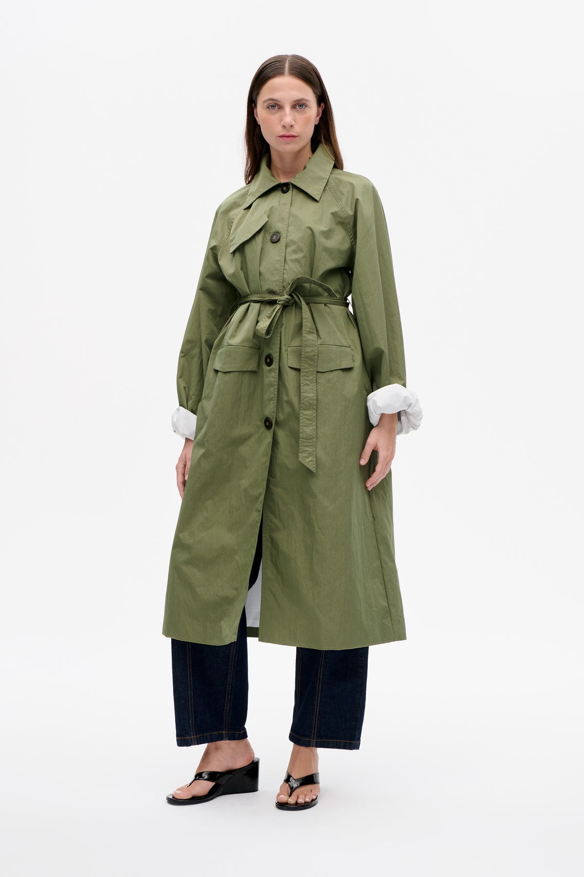 Front view of an olive green belted trench coat with a pointed collar, dark buttons and flap pockets, worn over dark jeans.