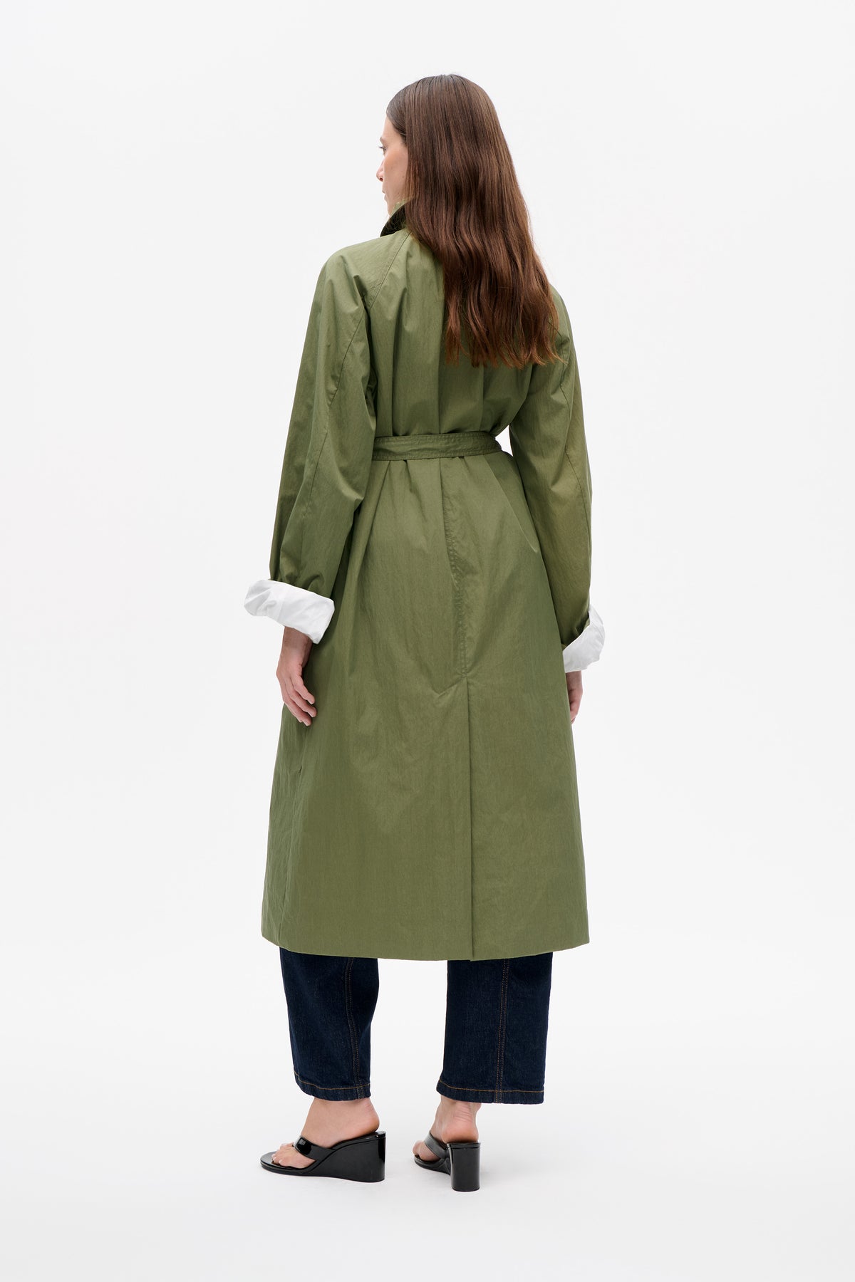 Rear view of a long olive-green belted coat with white turned-back cuffs, worn over dark jeans and black platform sandals.