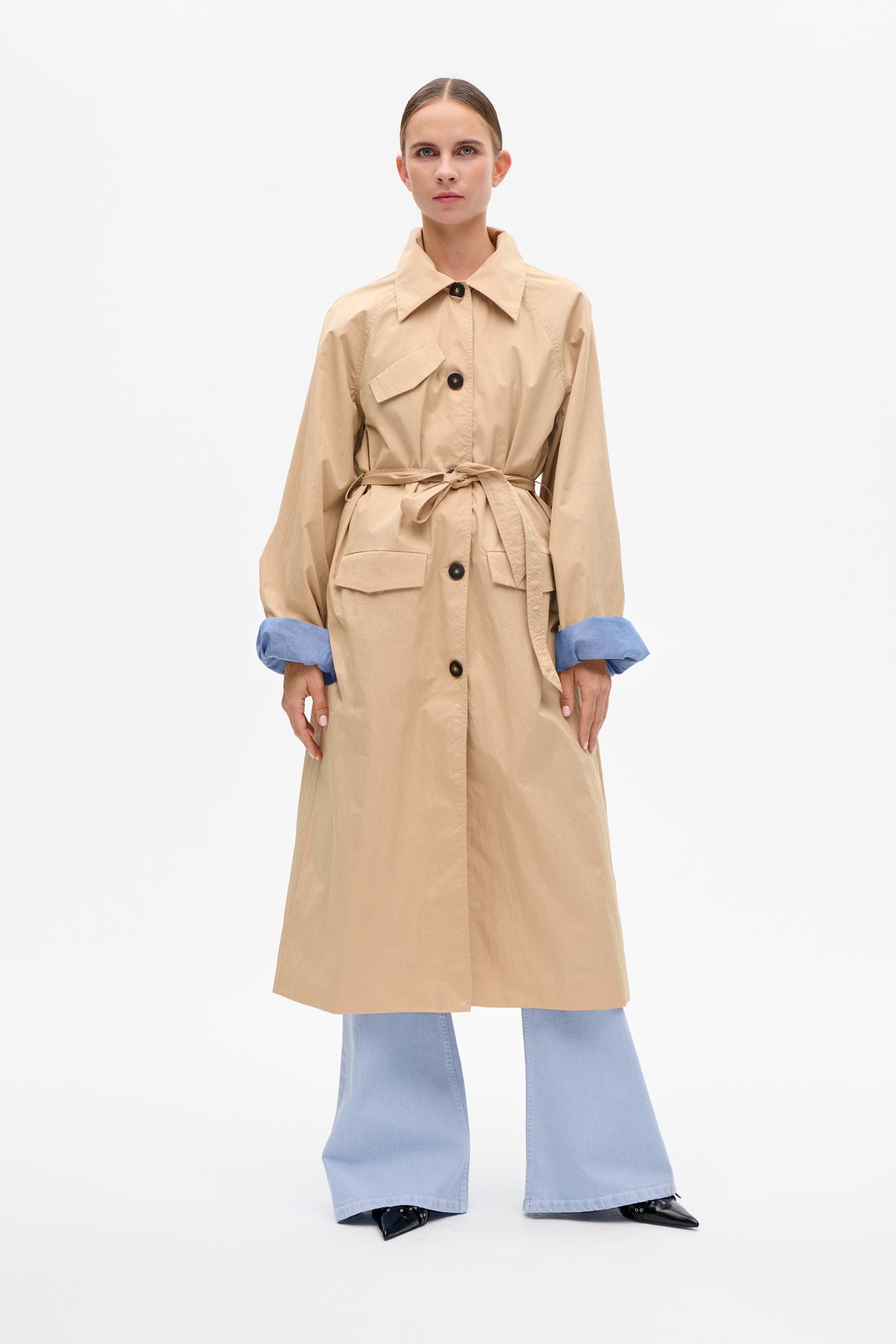 Front view of a beige belted trench coat with oversized collar, black buttons and flap pockets, styled with pale blue wide-leg trousers.