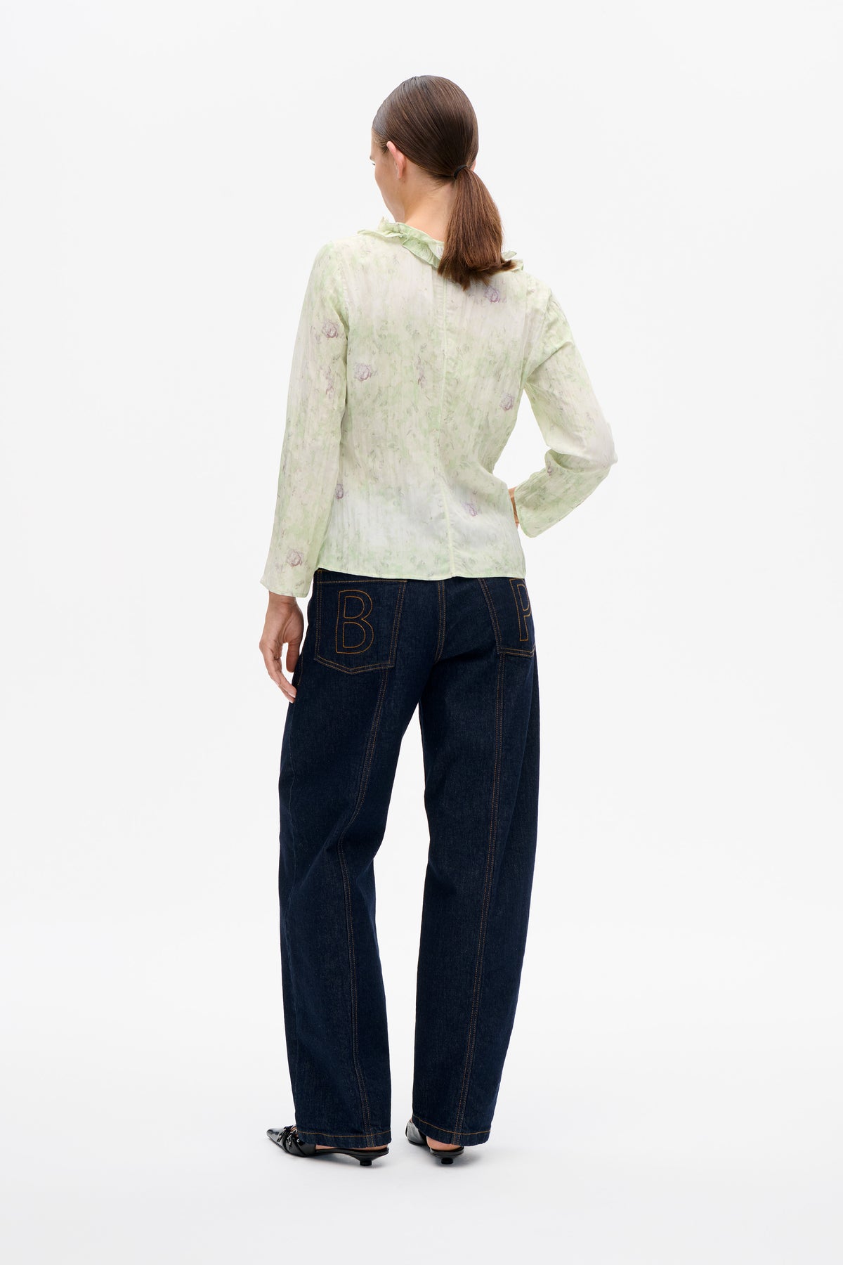 Achteraanzicht van een model dat een lichtgroene, bloemige blouse met lange mouwen en een kraag met ruches draagt, gecombineerd met donkere, wijdvallende jeans.