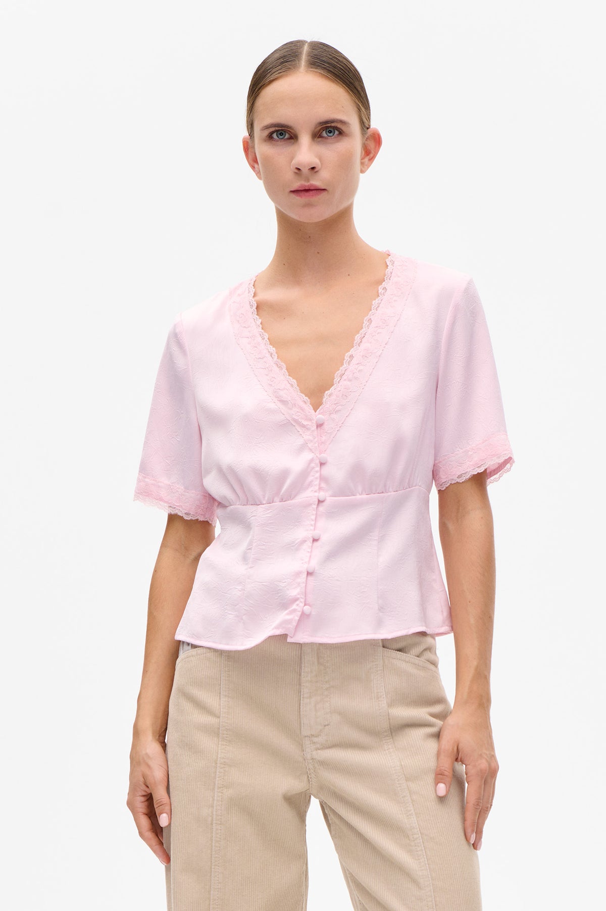 Front view of a pale pink short-sleeve blouse with a lace-trimmed V-neck, covered buttons and textured fabric.