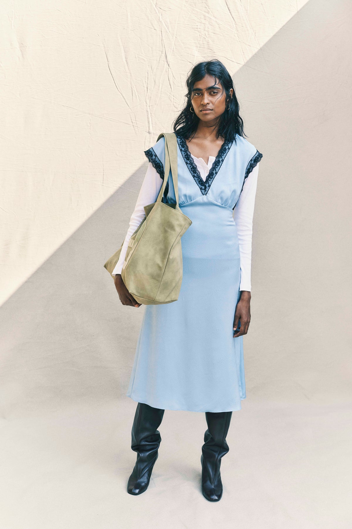 Campaign portrait of a model wearing a pale blue V-neck dress with black lace trim over a white shirt, carrying a khaki tote.