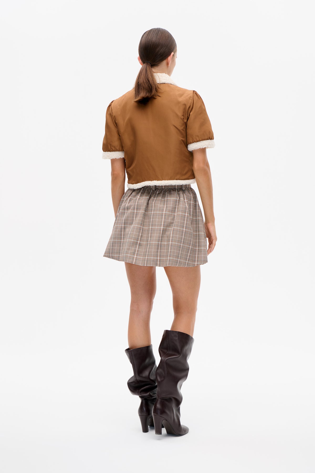 Back view of a rust-brown short-sleeve top with cream textured trim at the collar, cuffs and hem, styled with a plaid skirt.