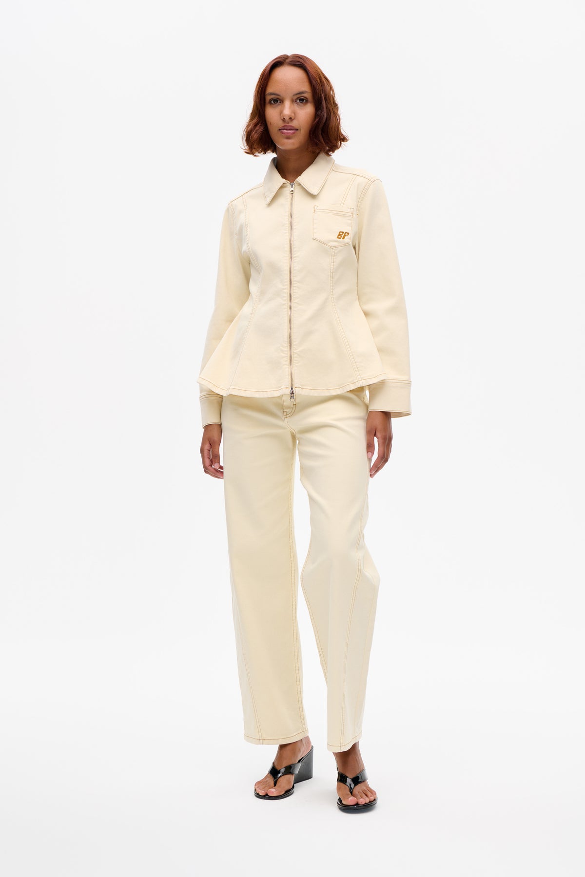 Front view of a model wearing a cream zip-front jacket with a pointed collar, chest pocket and brown BP embroidery, with matching trousers.
