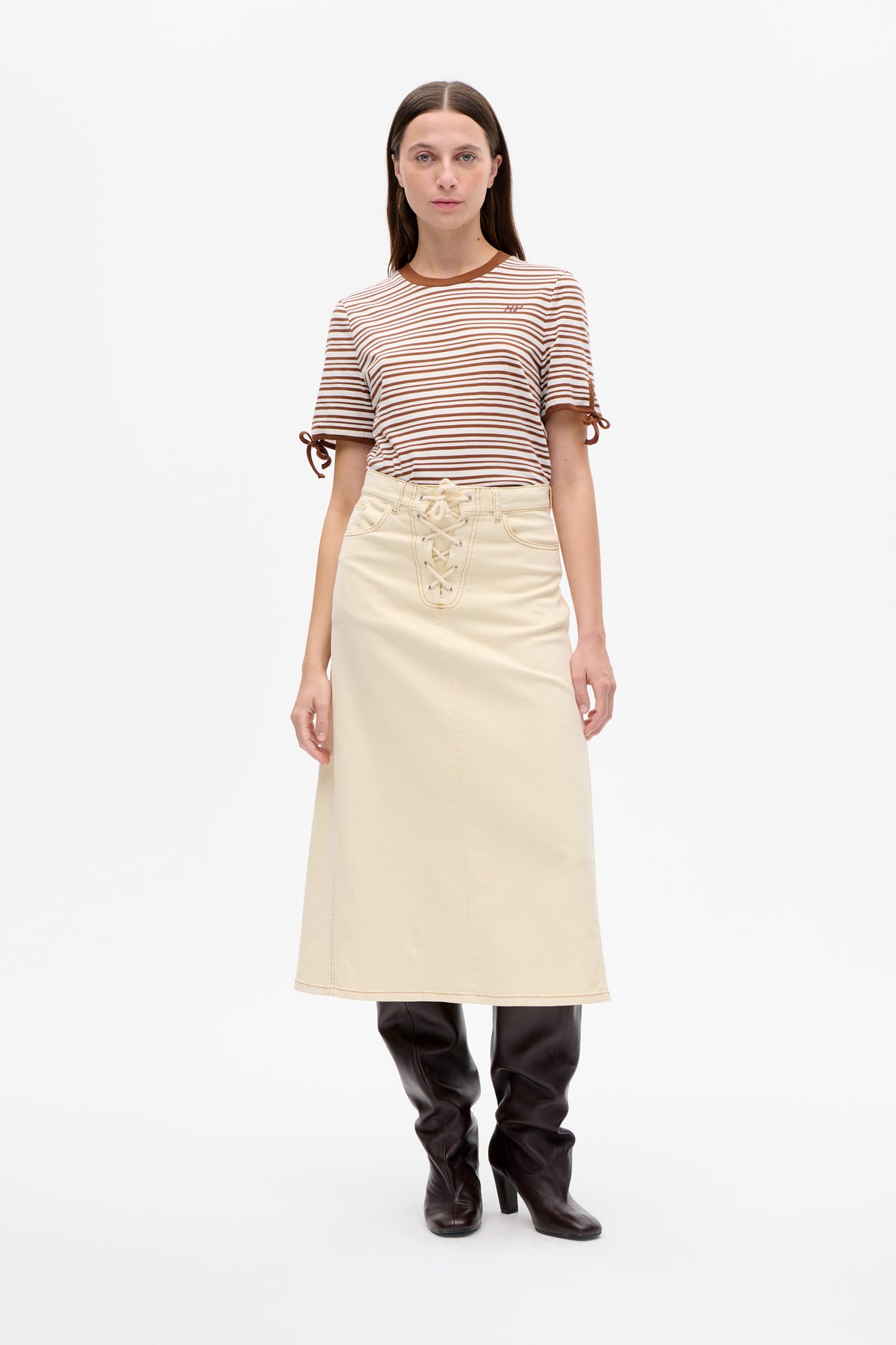 Frontalansicht eines Models mit weißem T‑Shirt mit braunen Streifen, braunem Rundhalsausschnitt und Bindebändern an den Ärmeln, kombiniert mit einem cremefarbenen Schnürrock.