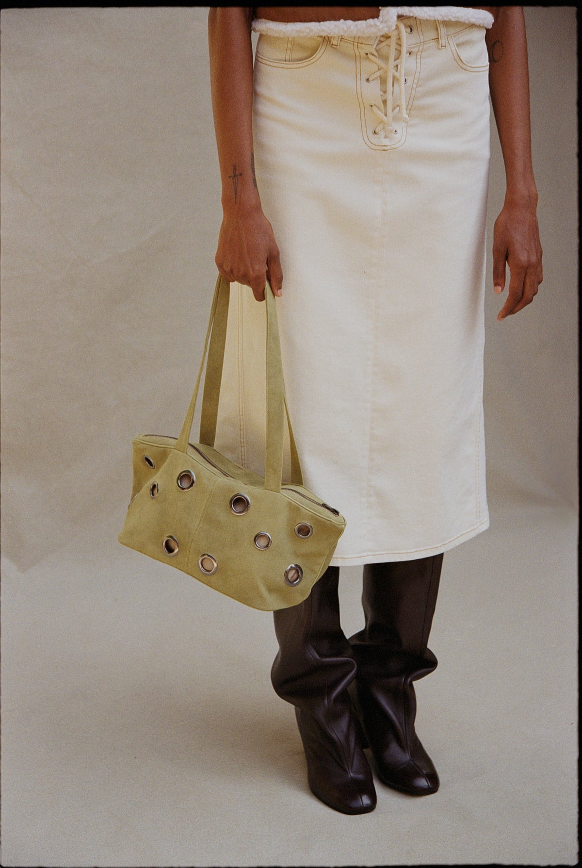 Cropped campaign view of a model in a cream lace-up skirt and fuzzy white top, carrying an olive eyelet tote and wearing black slouch boots.