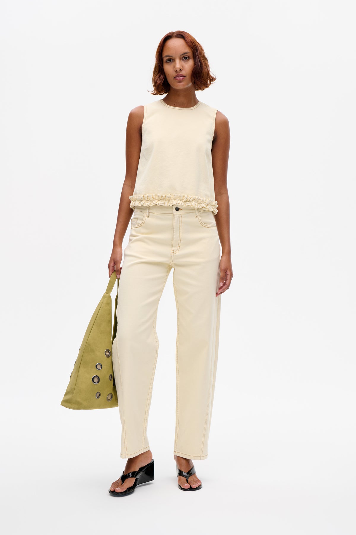 Model trägt ein cremefarbenes, ärmelloses Top mit Rüschensaum und passende, verkürzte Jeans mit weitem Bein; dazu eine olivgrüne Tote.