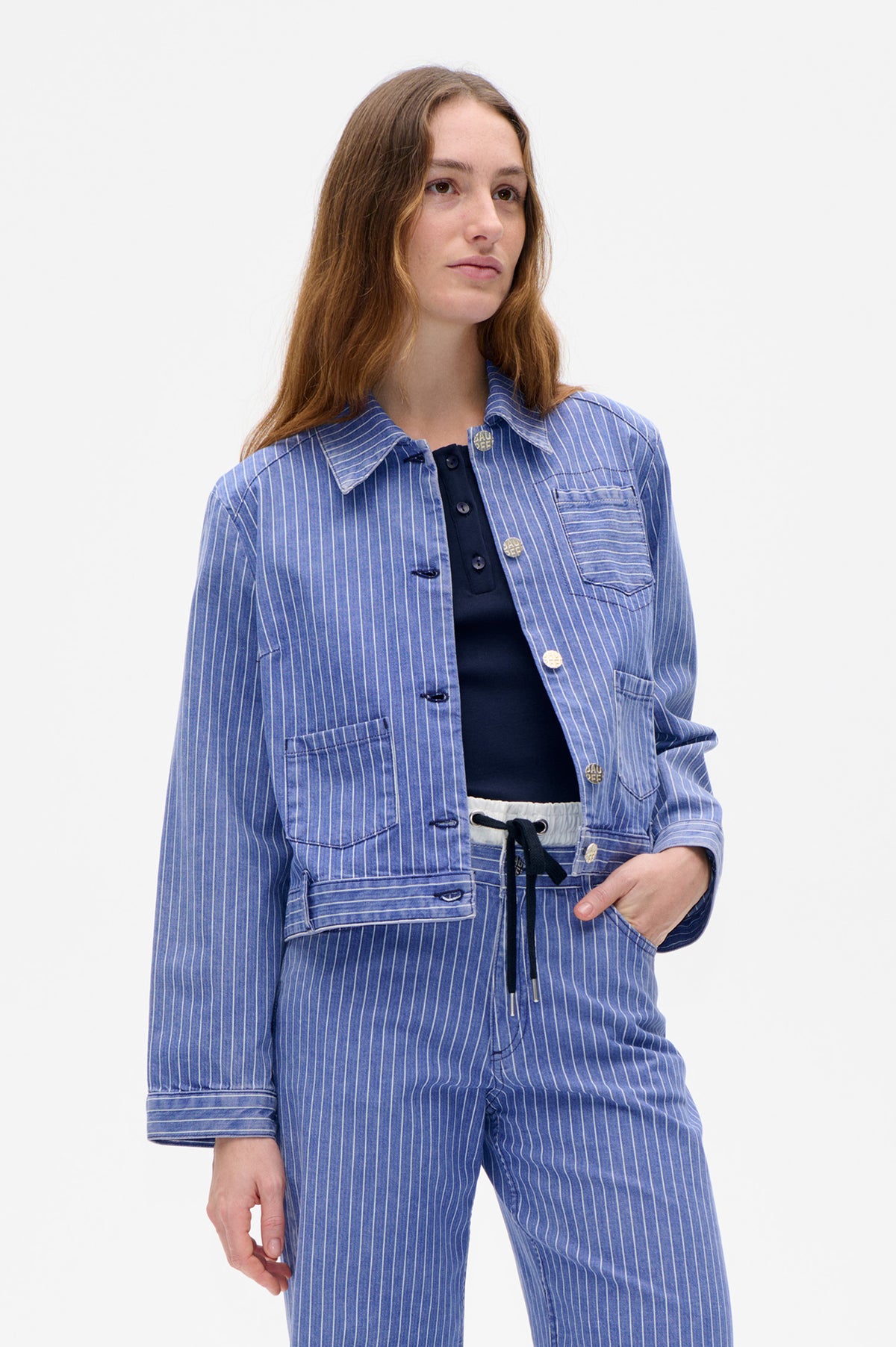 Model wearing a blue-and-white pinstriped utility jacket with patch pockets over a navy top and matching trousers.