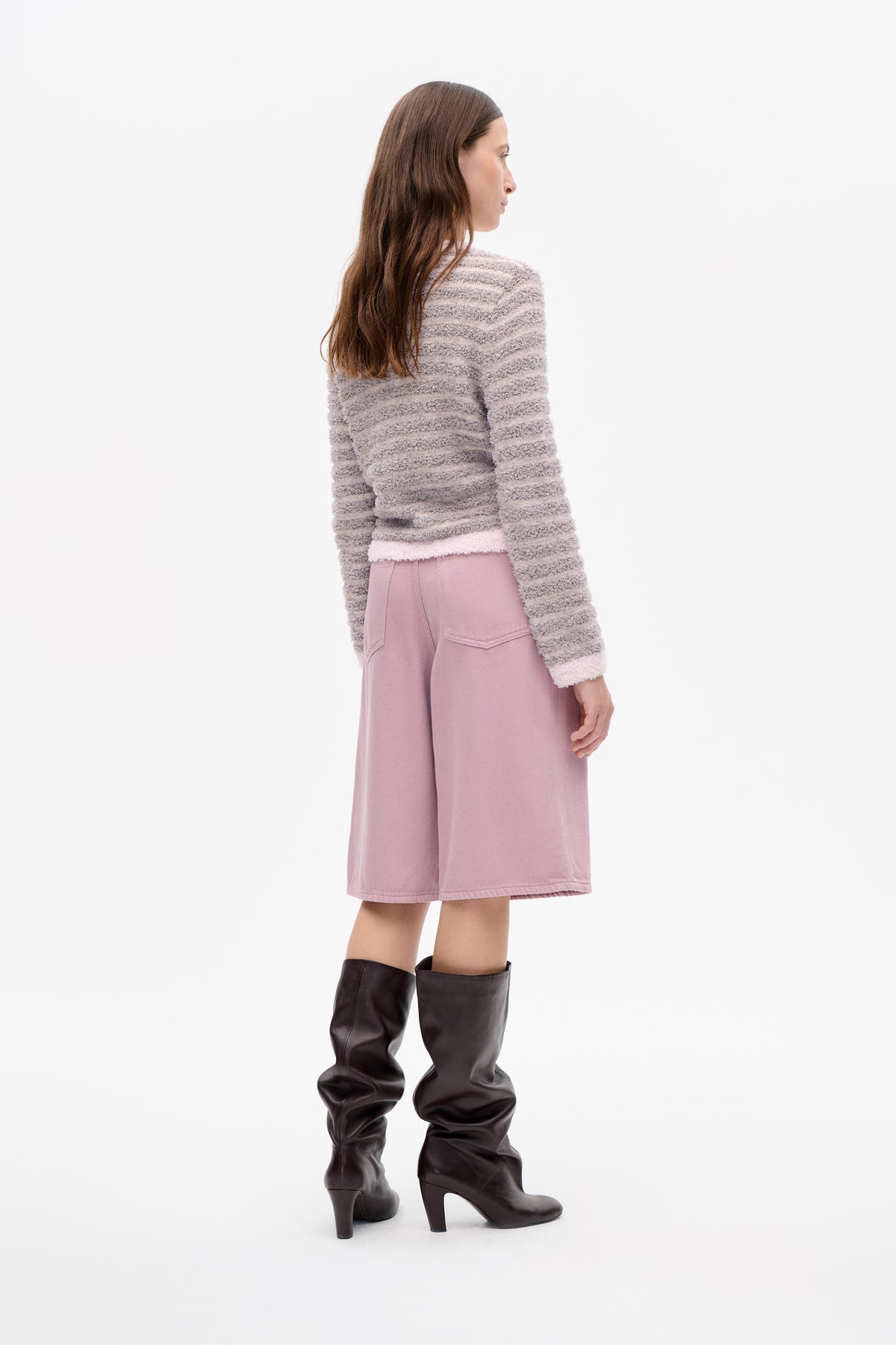 Side view of a model wearing a textured cream and grey striped sweater, dusty pink knee-length skirt and dark knee-high heeled boots.
