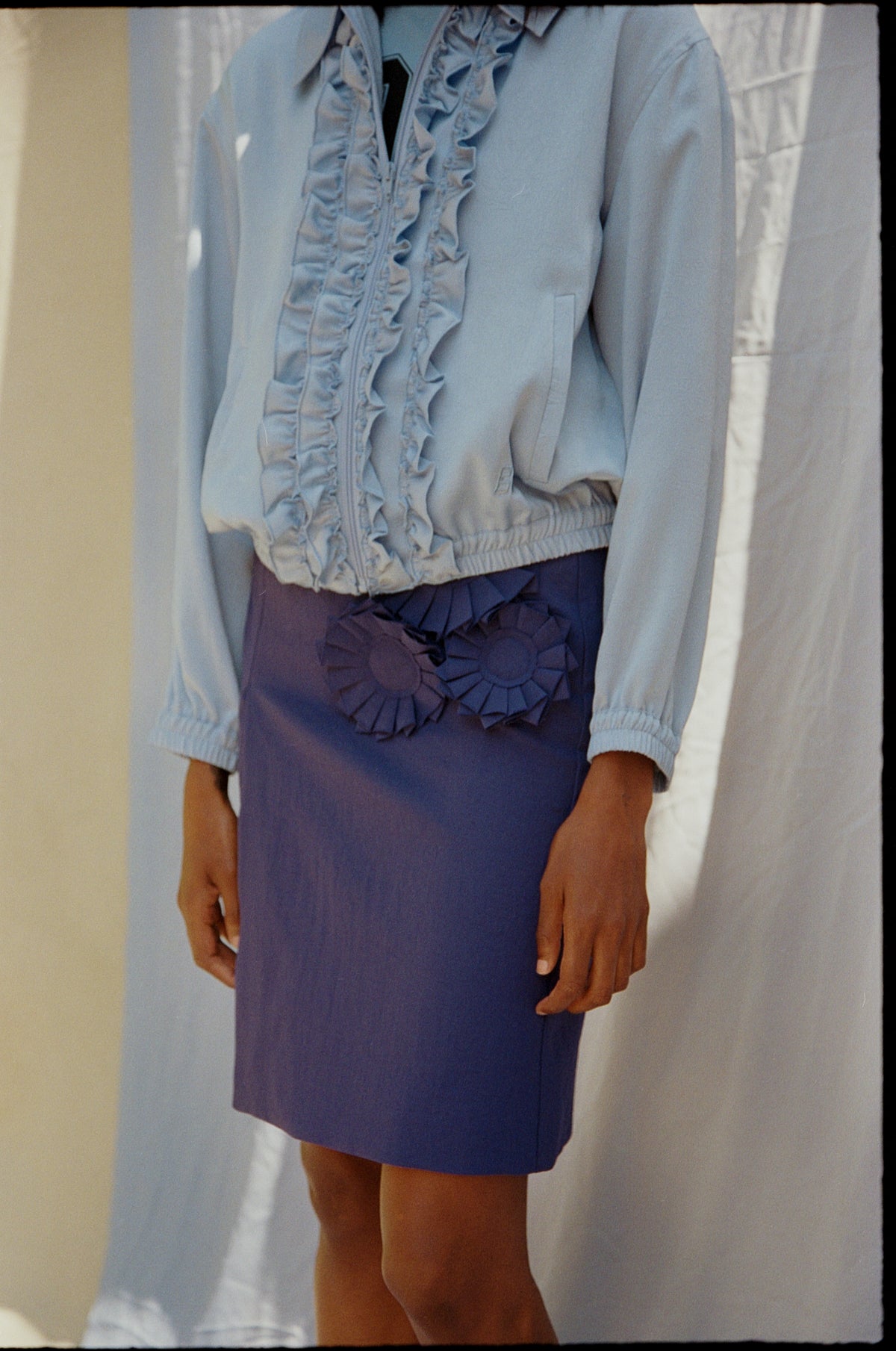Cropped campaign view of a pale blue jacket over a ruffled light blue blouse and a deep blue dress with pleated detailing.