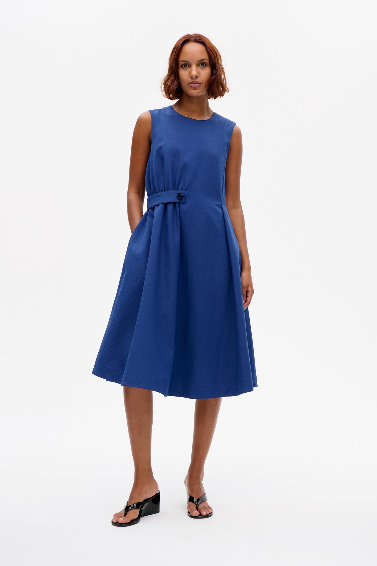 Model wearing a sleeveless blue dress with a round neckline, gathered waist and side buckle detail, shown from the front.