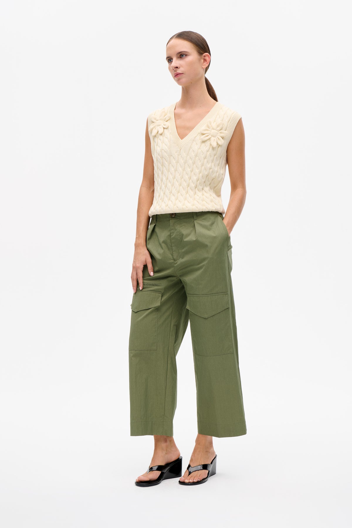 Model wearing an ivory cable-knit sleeveless V-neck sweater vest with raised flower details and olive wide-leg cargo trousers.