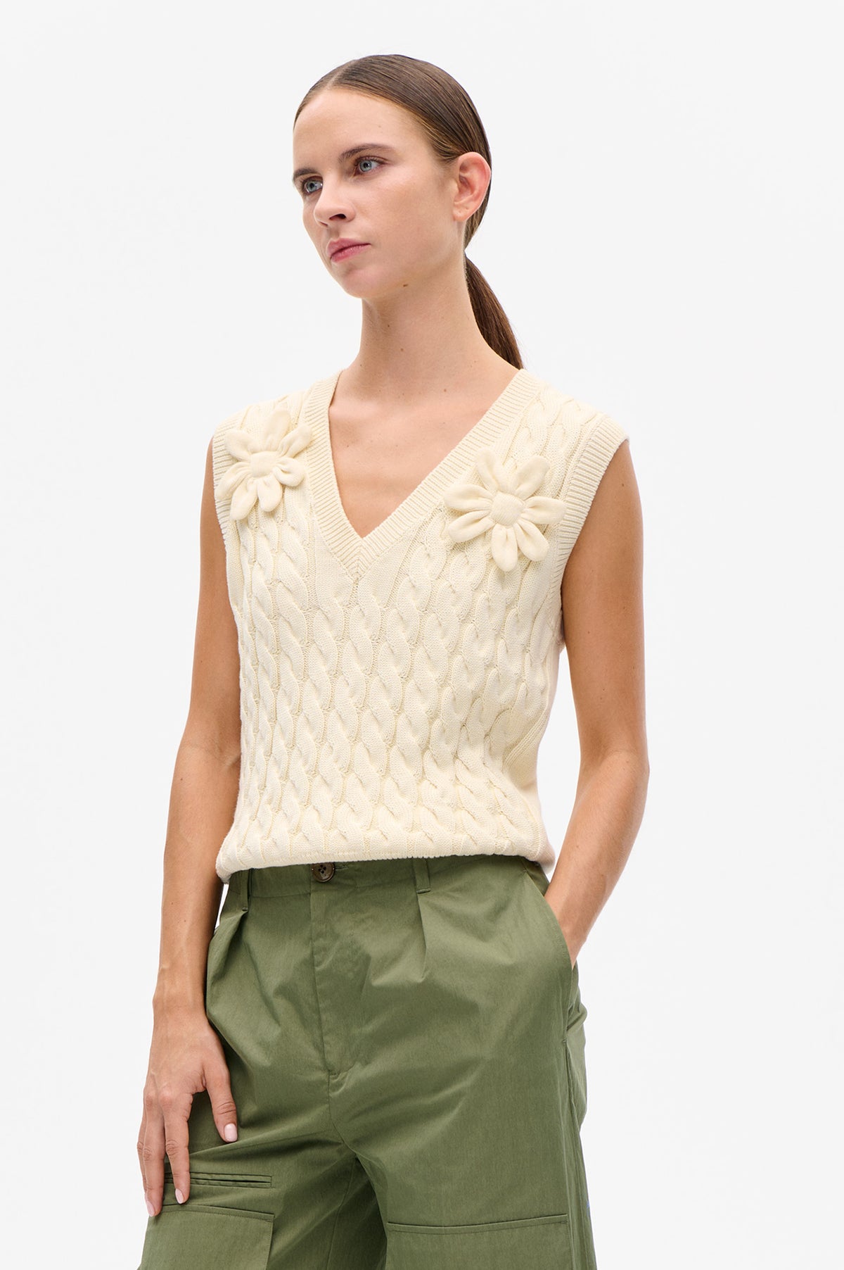 Model wearing a cream sleeveless cable-knit V-neck vest with raised flower details and olive green trousers.