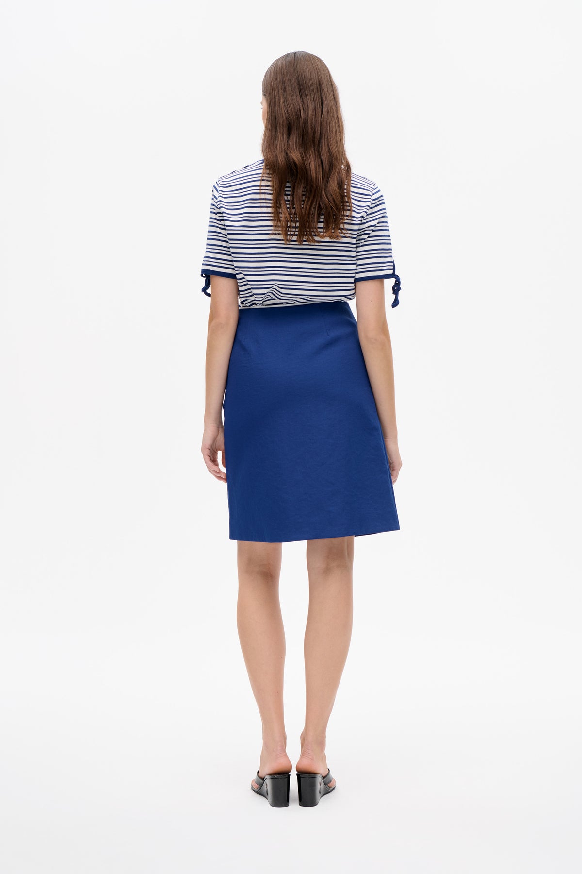 Rückenansicht eines Models, das ein weißes T-Shirt mit marineblauen Querstreifen und Schleifen an den Ärmeln trägt, kombiniert mit einem einfarbigen, knielangen blauen Rock.