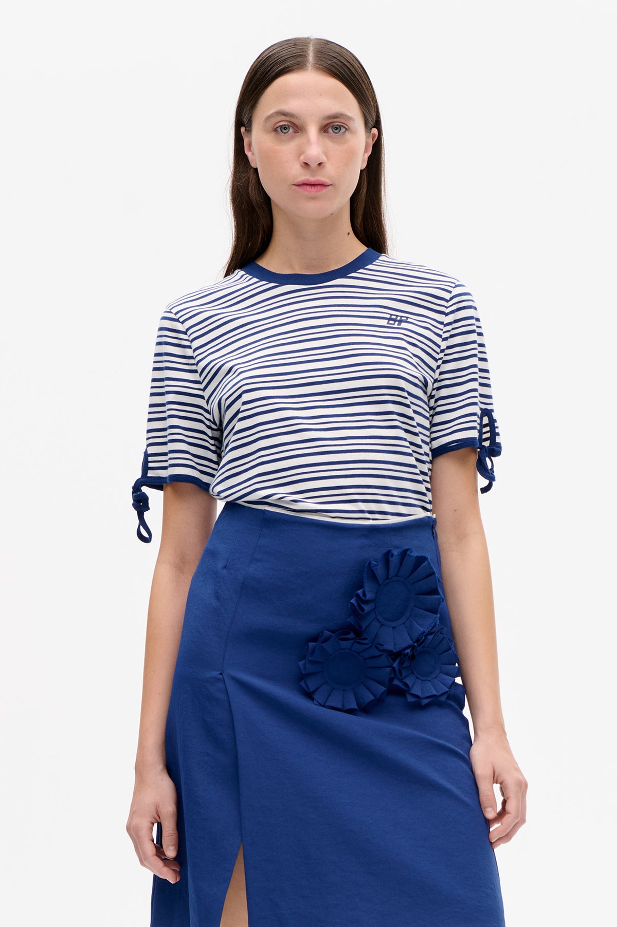 Front view of a model wearing a white T-shirt with navy stripes and sleeve ties, paired with a cobalt blue skirt with ruffled flower appliqués.