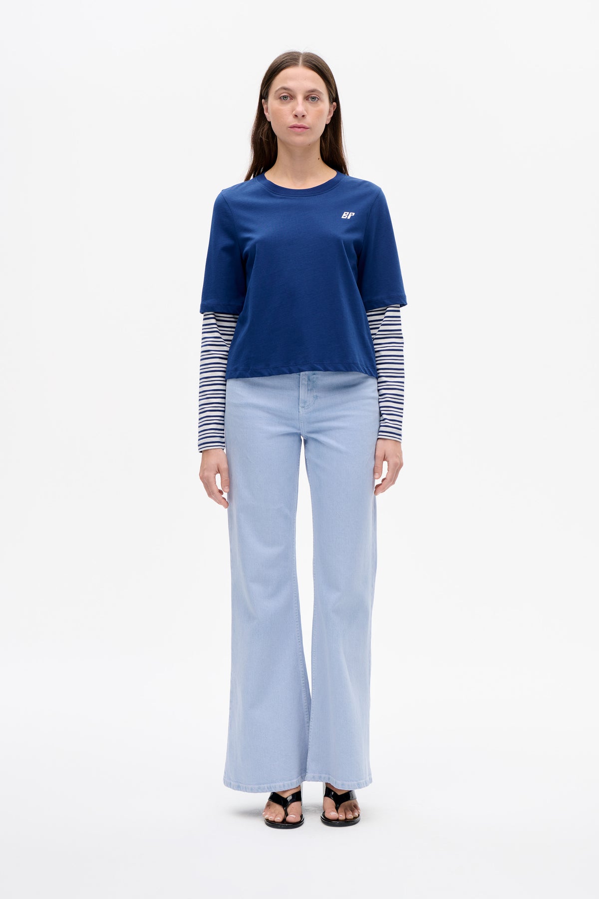 Front view of a model wearing a blue short-sleeve T-shirt with a small white BP logo over a navy-and-white striped long-sleeve top, light-wash wide-leg jeans and black sandals.