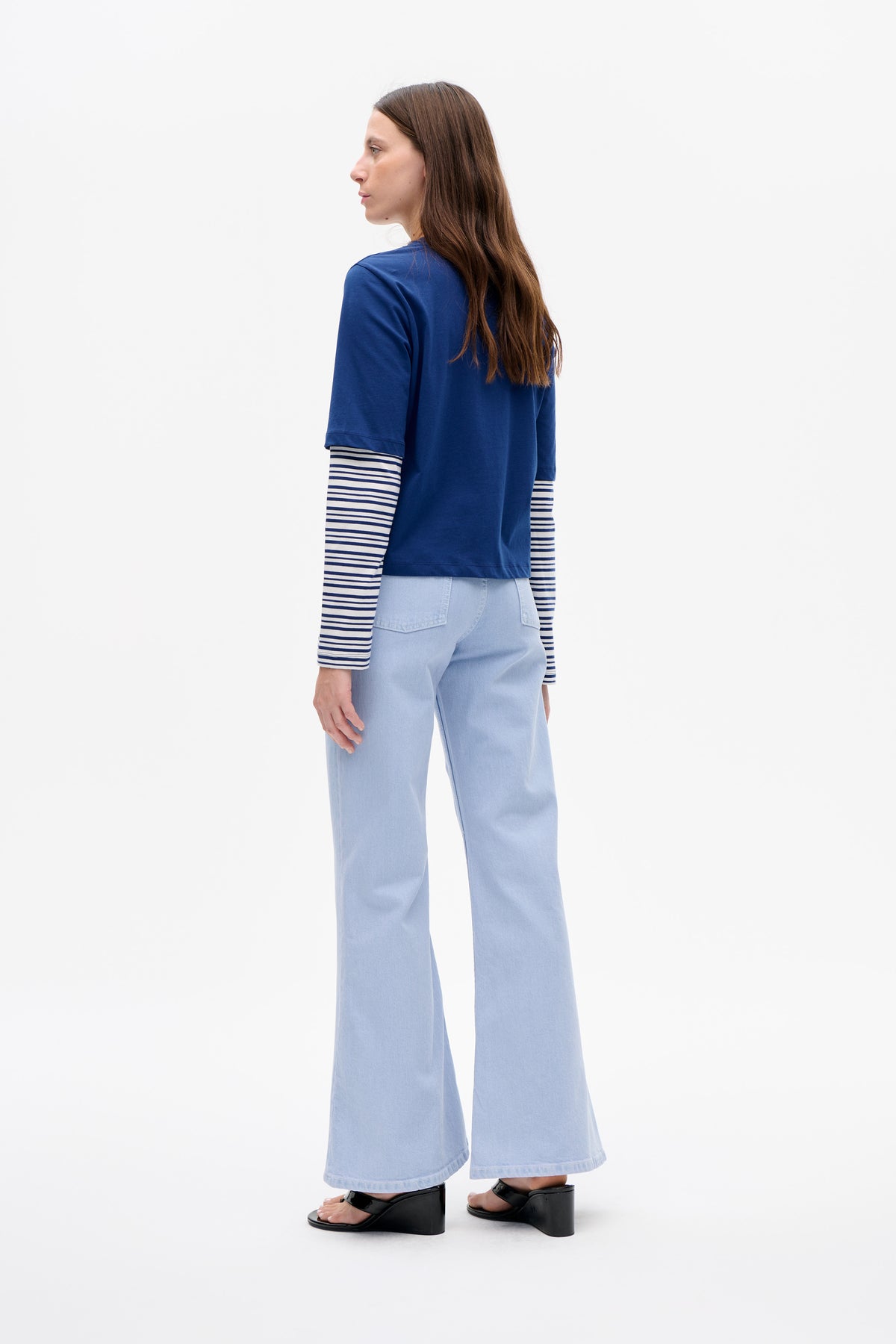 Model in side-back view wearing a cobalt blue short-sleeve T-shirt over a navy-and-white striped long-sleeve top and pale blue wide-leg jeans.