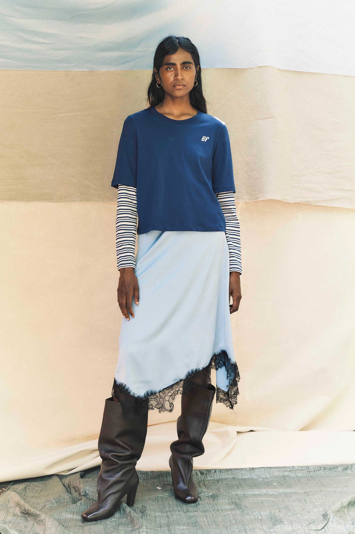 Model wearing a blue short-sleeved T-shirt with a small BP chest logo over a striped long-sleeved top, pale blue skirt with black lace trim, and tall dark boots.