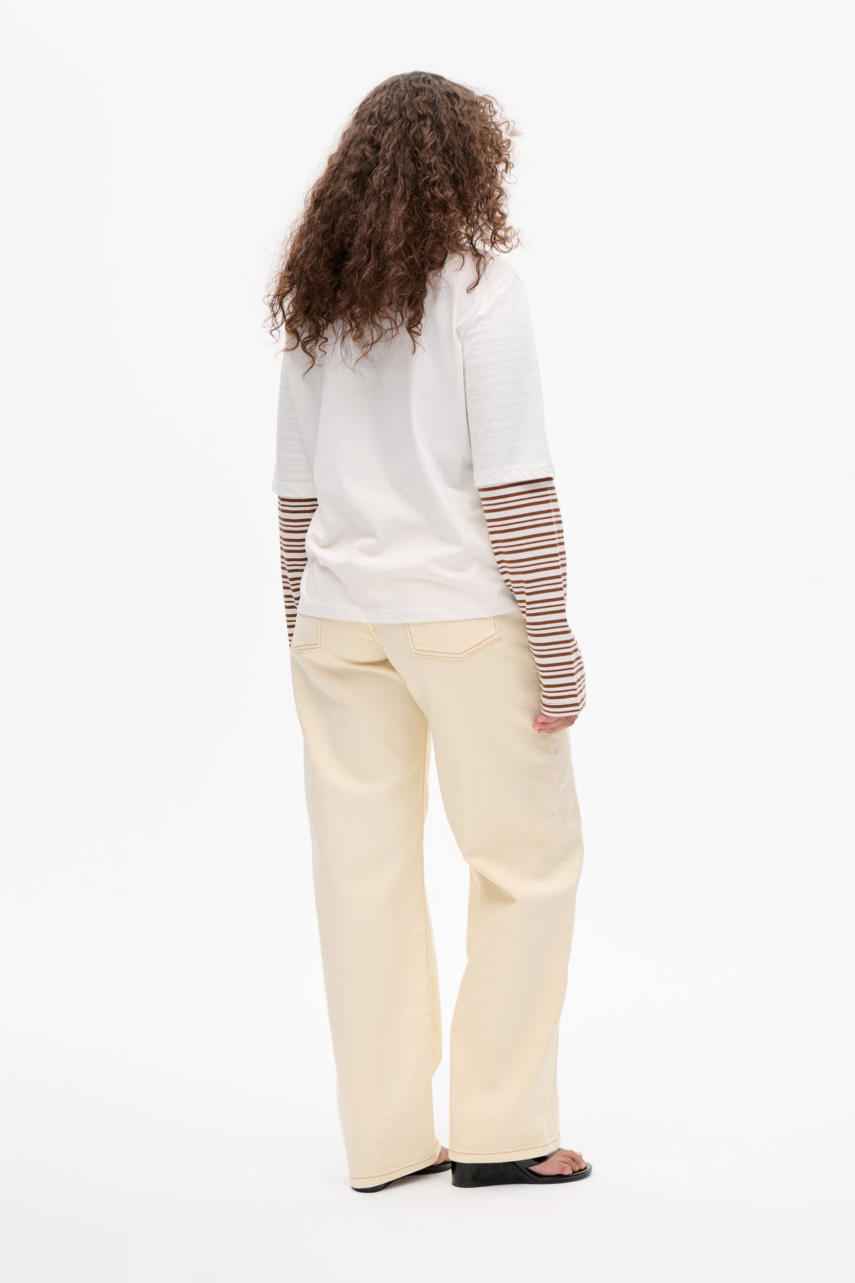 Vue de dos d'un mannequin portant un t-shirt blanc à manches courtes superposé à un top à manches longues rayé marron et blanc, associé à un pantalon large couleur crème.