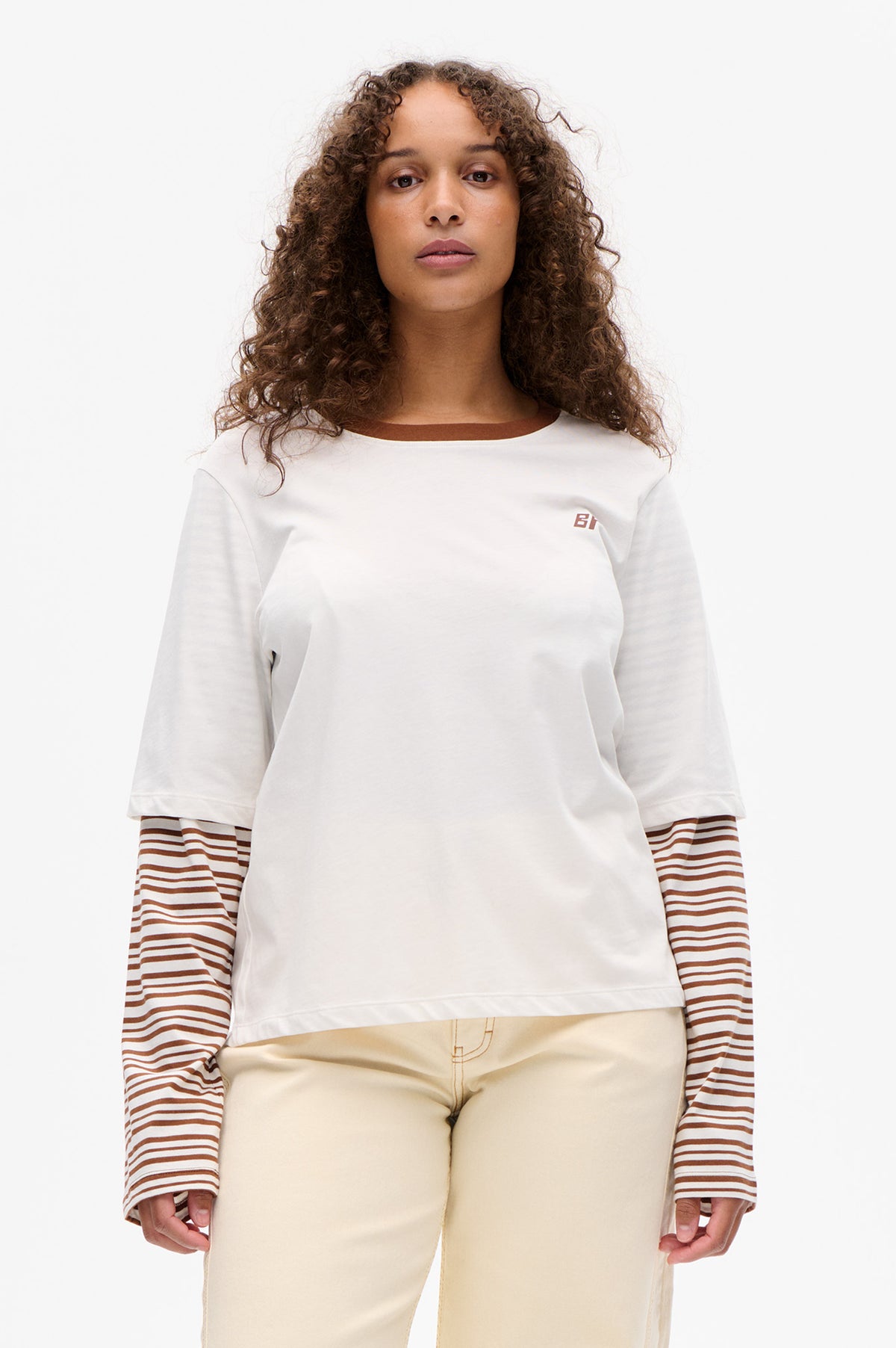Front view of a white short-sleeved top with a small brown chest logo layered over a brown-and-white striped long-sleeved shirt, paired with cream trousers.