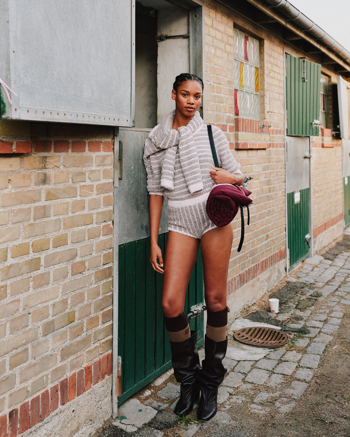 Mannequin à côté d'un bâtiment en briques portant un haut texturé gris pâle avec un col drapé, un short assorti, des bottes hautes noires et un sac bordeaux.