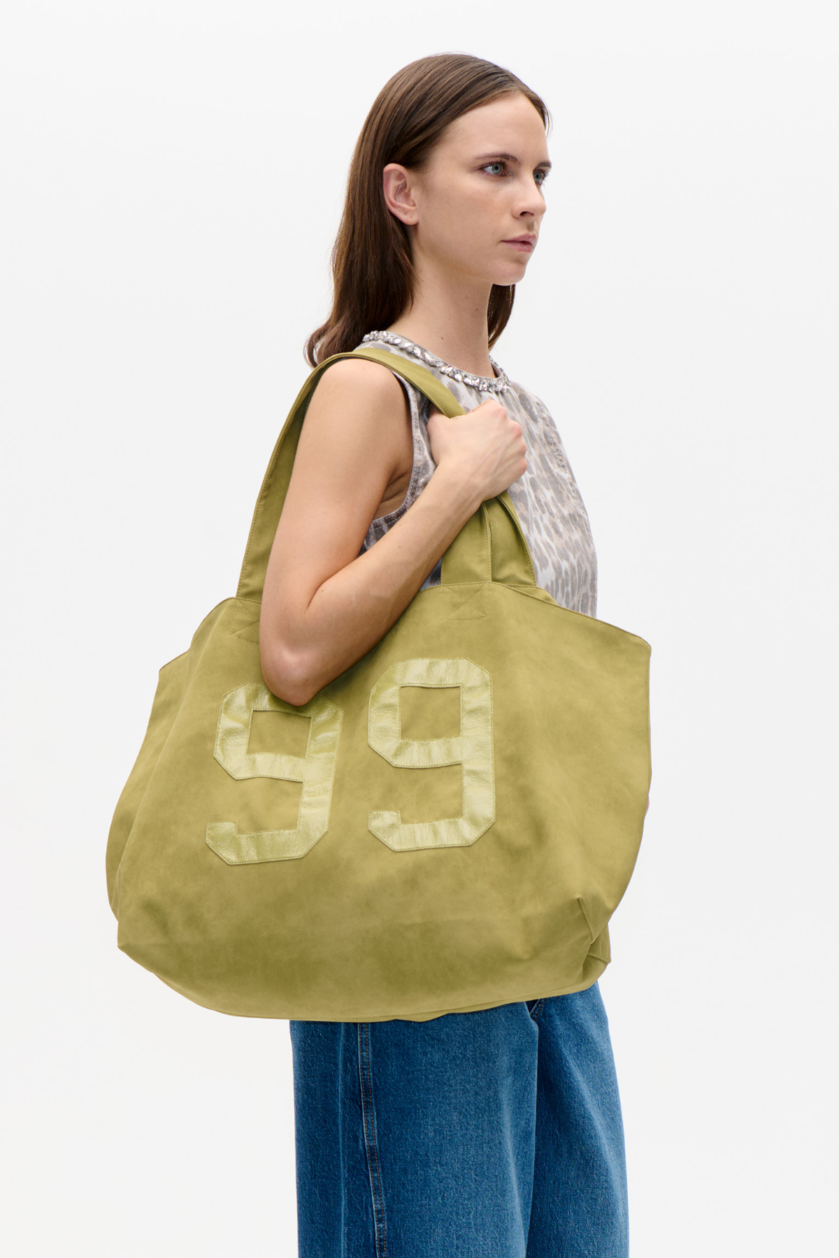 Model carrying an oversized olive-green tote with long handles and a large pale 99 appliqué, styled with a patterned sleeveless top and blue jeans.
