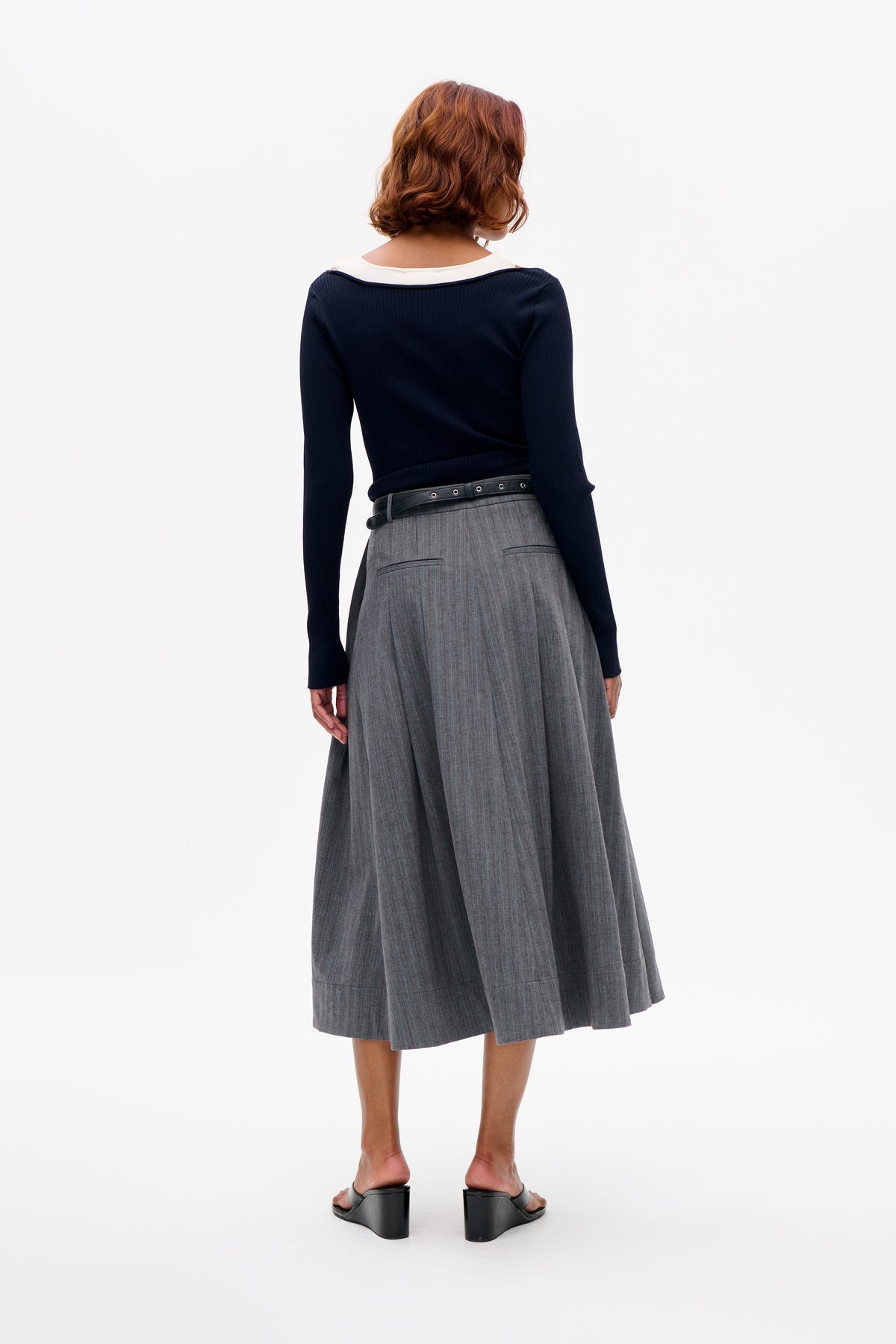 Back view of a navy ribbed long-sleeve top with a cream collar, paired with a grey pleated skirt and black eyelet belt.