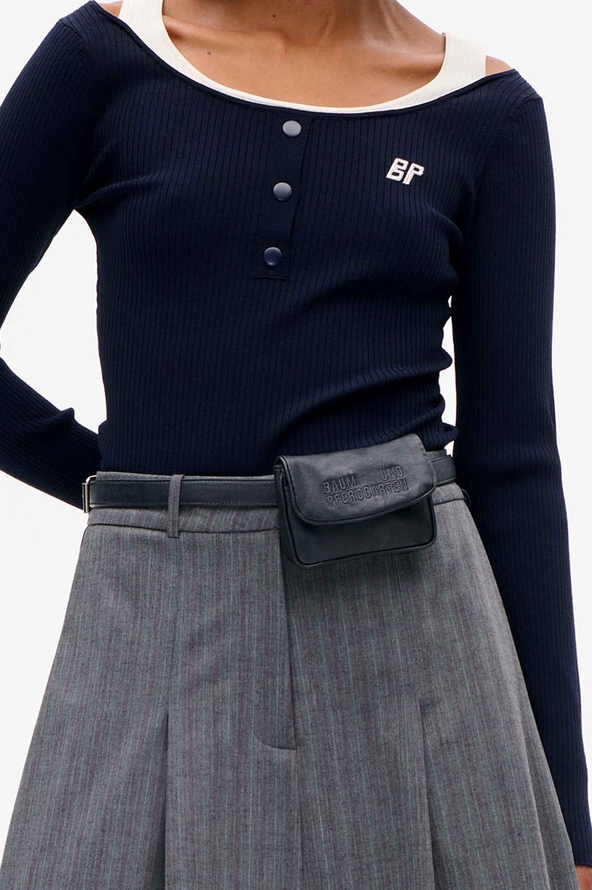 Close-up of a navy ribbed long-sleeve top with front buttons and embroidered initials, layered over a white tank; grey trousers and a black belt bag are visible.