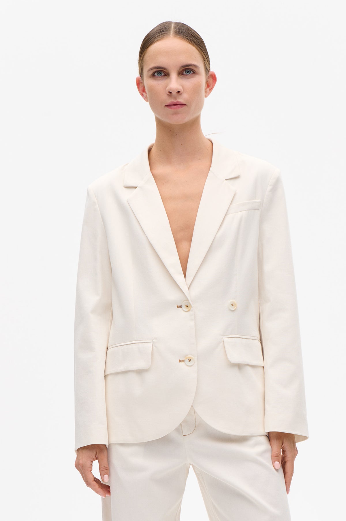 Front view of an ivory double-breasted blazer with notched lapels, light buttons and flap pockets, worn with matching trousers.