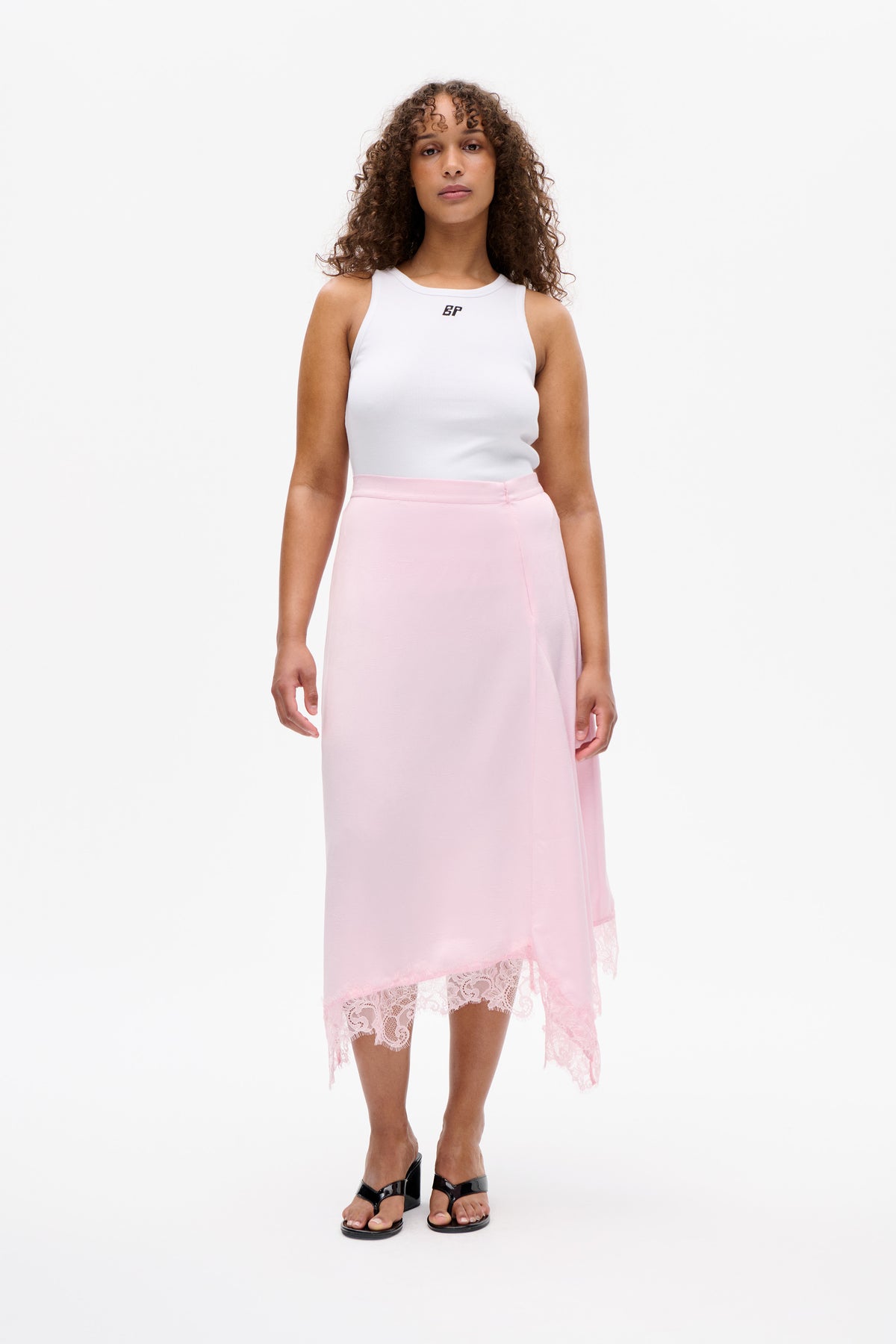 Front view of a model wearing a white sleeveless top and pale pink midi skirt with an asymmetric lace-trimmed hem.
