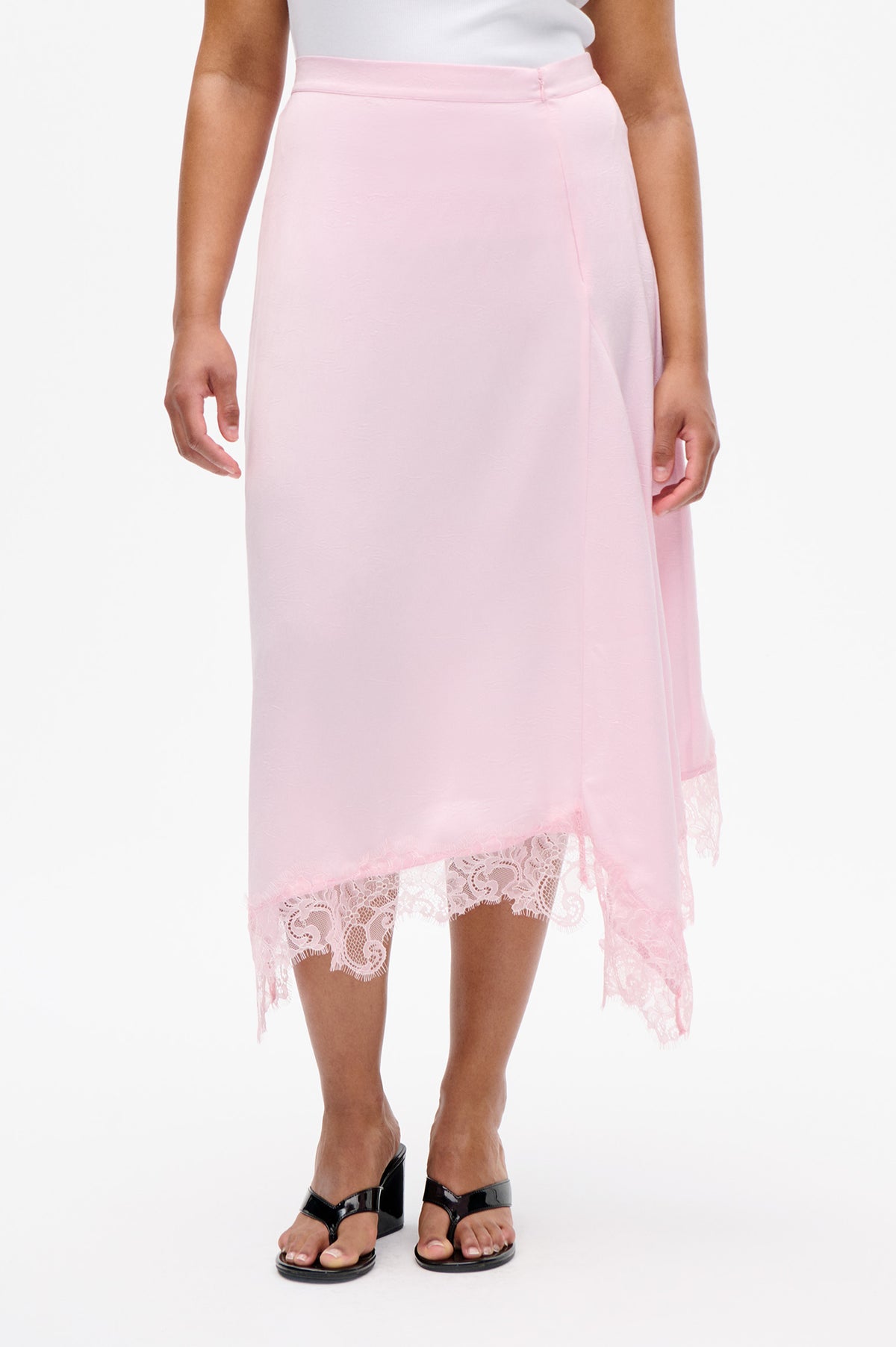 Front detail of a pale pink midi skirt with a high waistband and asymmetric floral lace hem, worn with a white top.