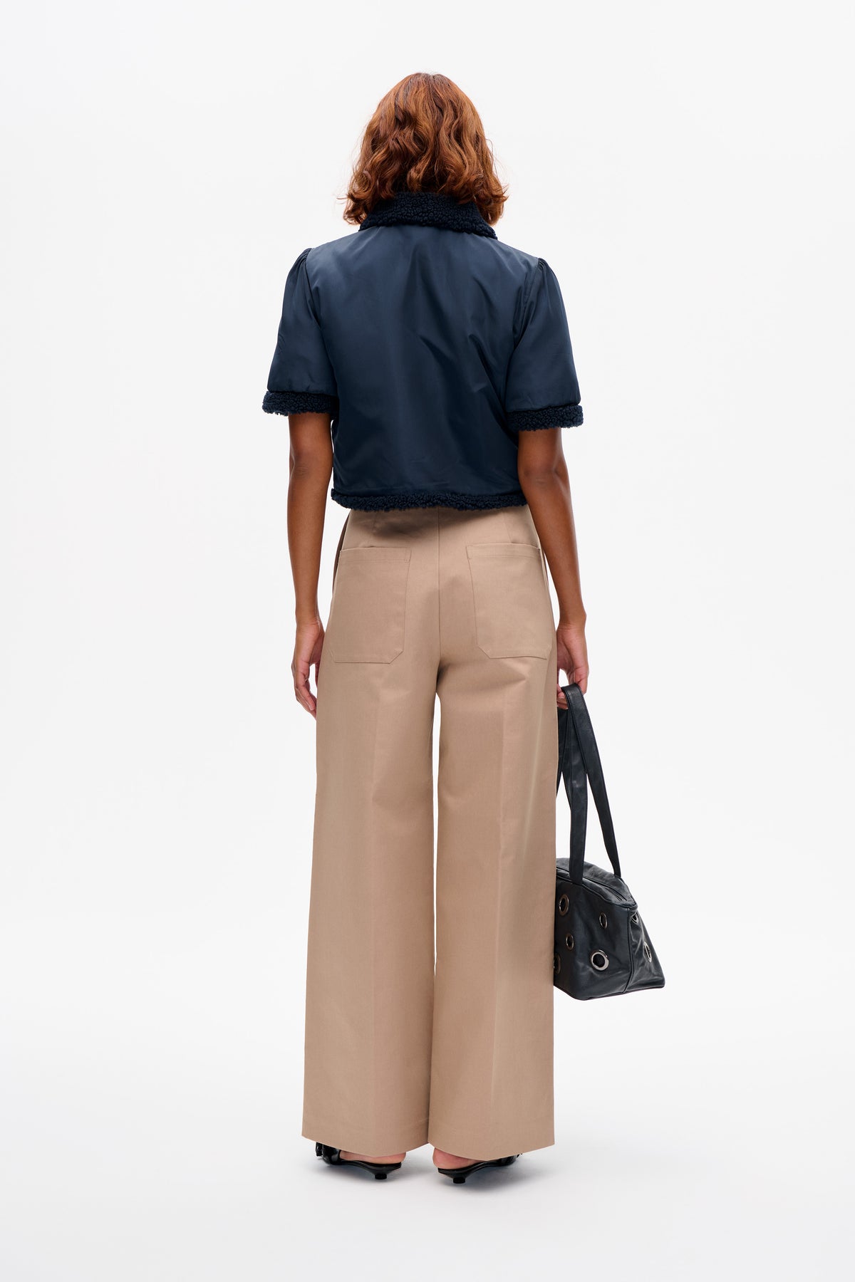 Rückenansicht eines Models, das eine marineblaue, kurzärmelige Jacke mit strukturiertem Besatz an Kragen, Bündchen und Saum sowie beige, weit geschnittene Hosen trägt.