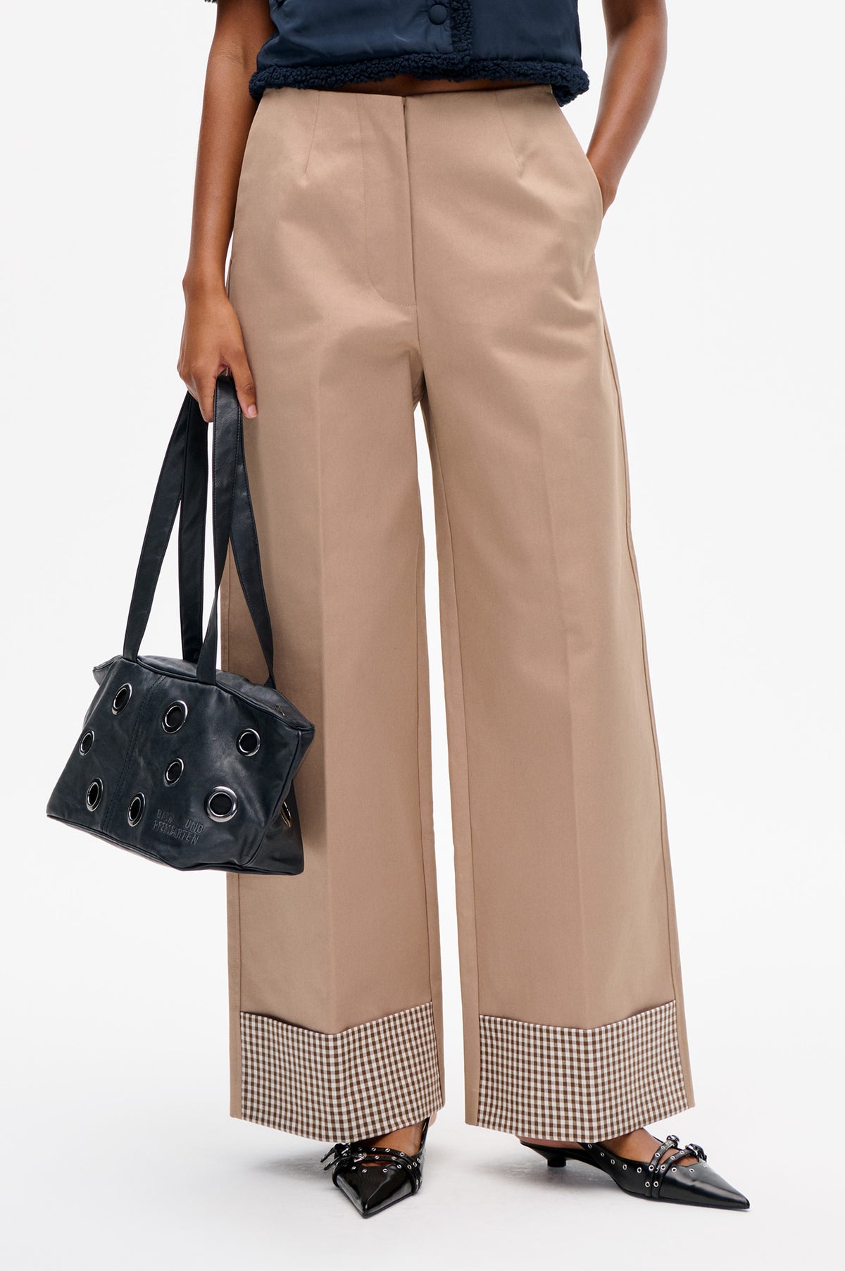 Nahaufnahme, Vorderansicht einer taupefarbenen Hose mit weitem Bein und braun-cremefarbenen Vichy-Karo-Einsätzen am Saum, gestylt mit einem navyblauen Top und einer schwarzen Tasche mit Ösen.