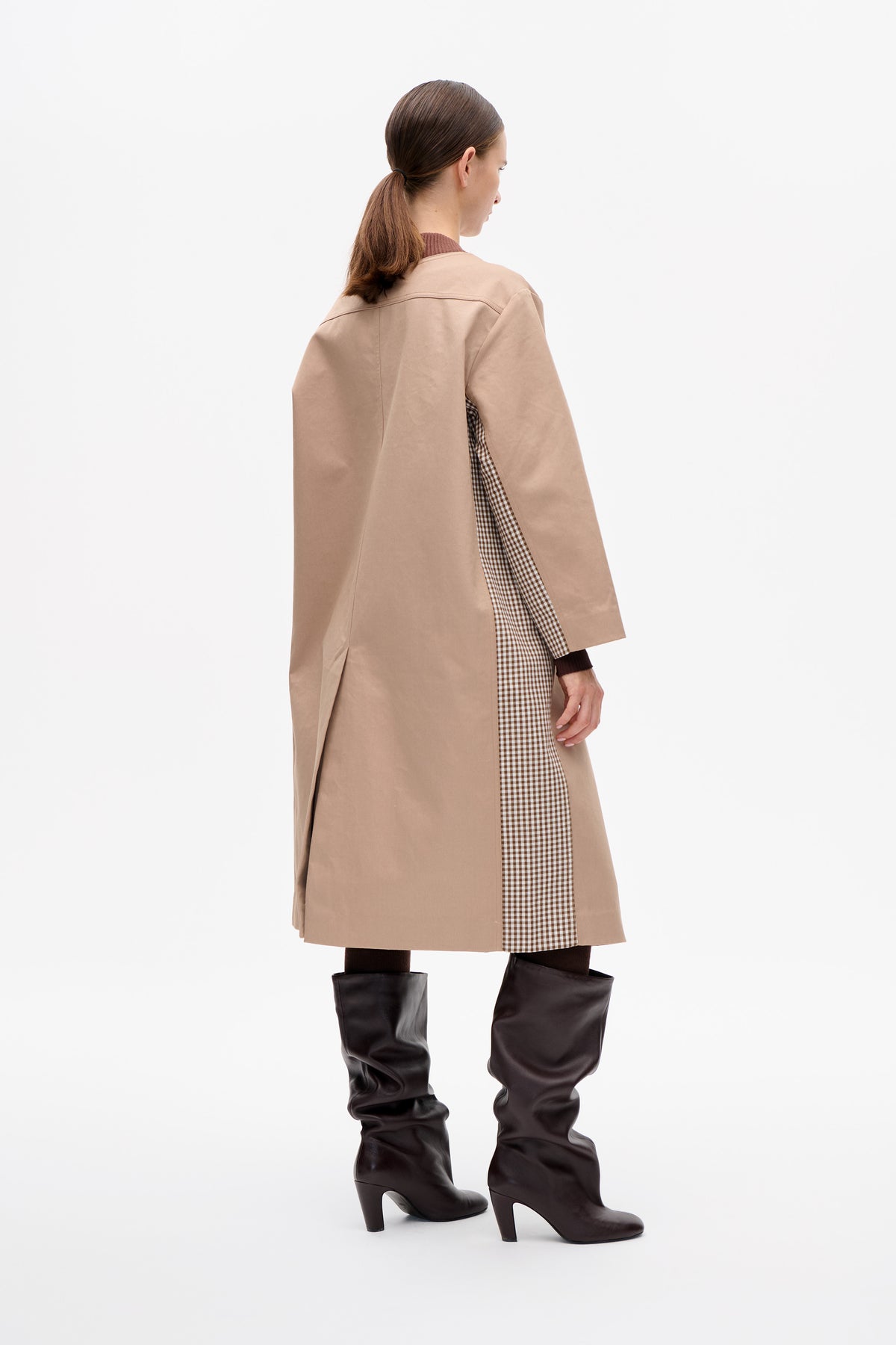Rear three-quarter view of a model in a long beige coat with a brown-and-cream checked panel, checked dress and dark heeled boots.