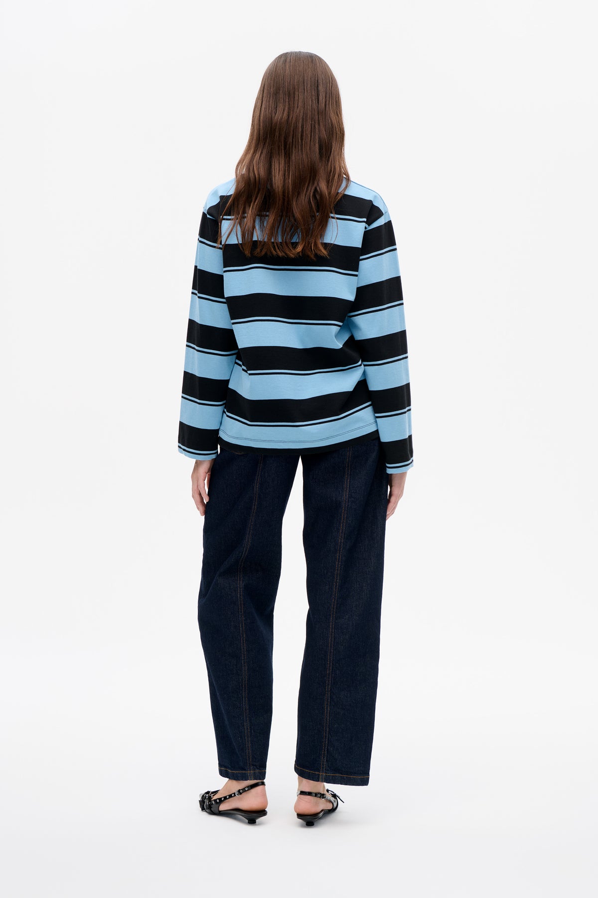 Back view of a model wearing a long-sleeved top with broad light blue and black horizontal stripes, dark jeans and black sandals.