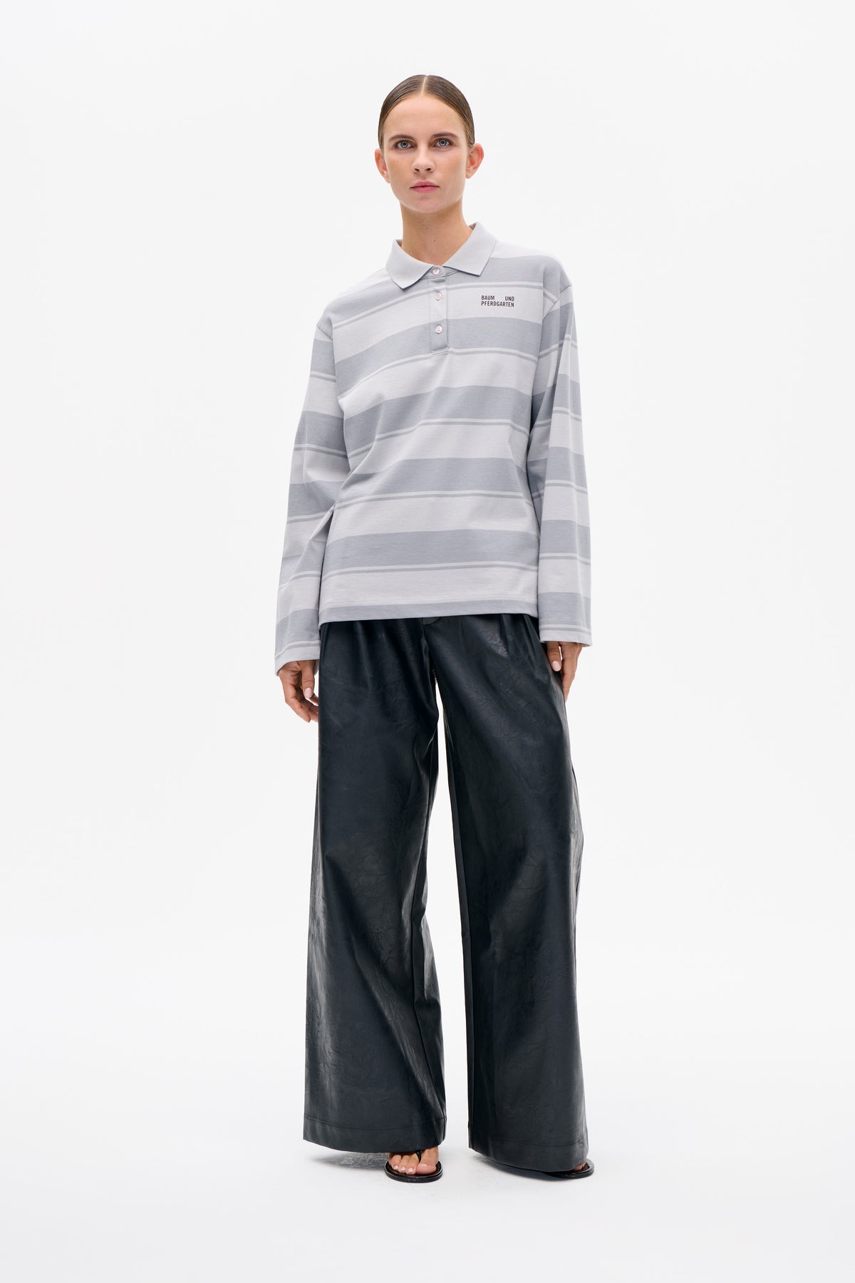 Front view of a model wearing a grey-and-white striped long-sleeve polo shirt with a small chest logo and black wide-leg trousers.
